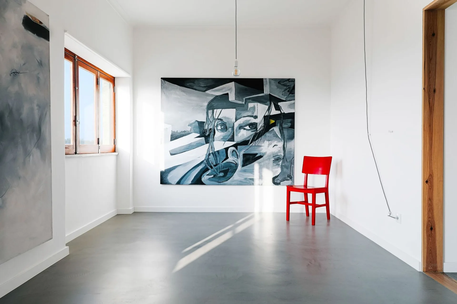A minimalist white room with a gray concrete floor, a large monochrome wall painting, and a single bright red wooden chair.