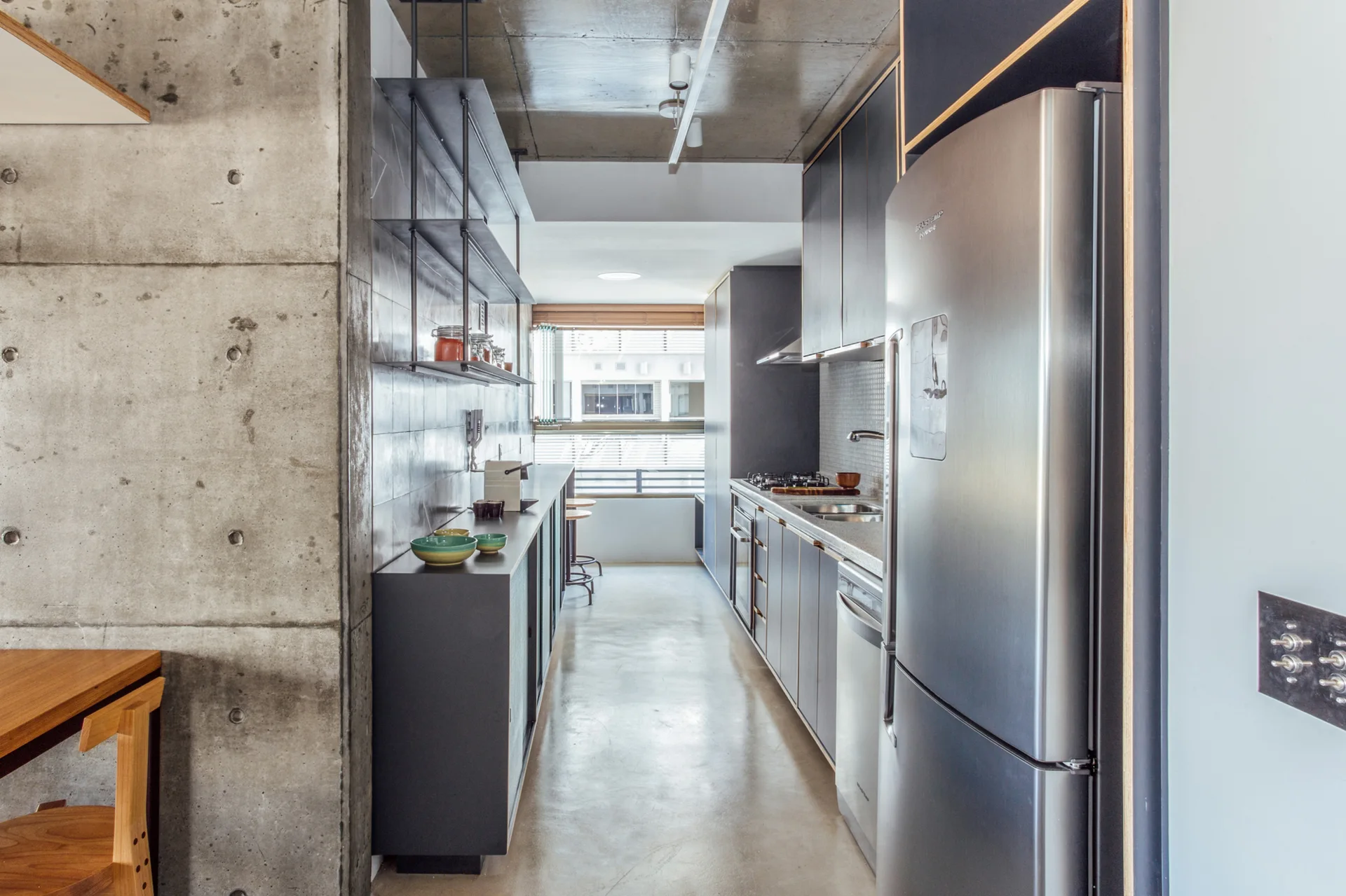 A narrow galley kitchen with raw exposed concrete walls, dark charcoal cabinetry, and a stainless steel refrigerator.