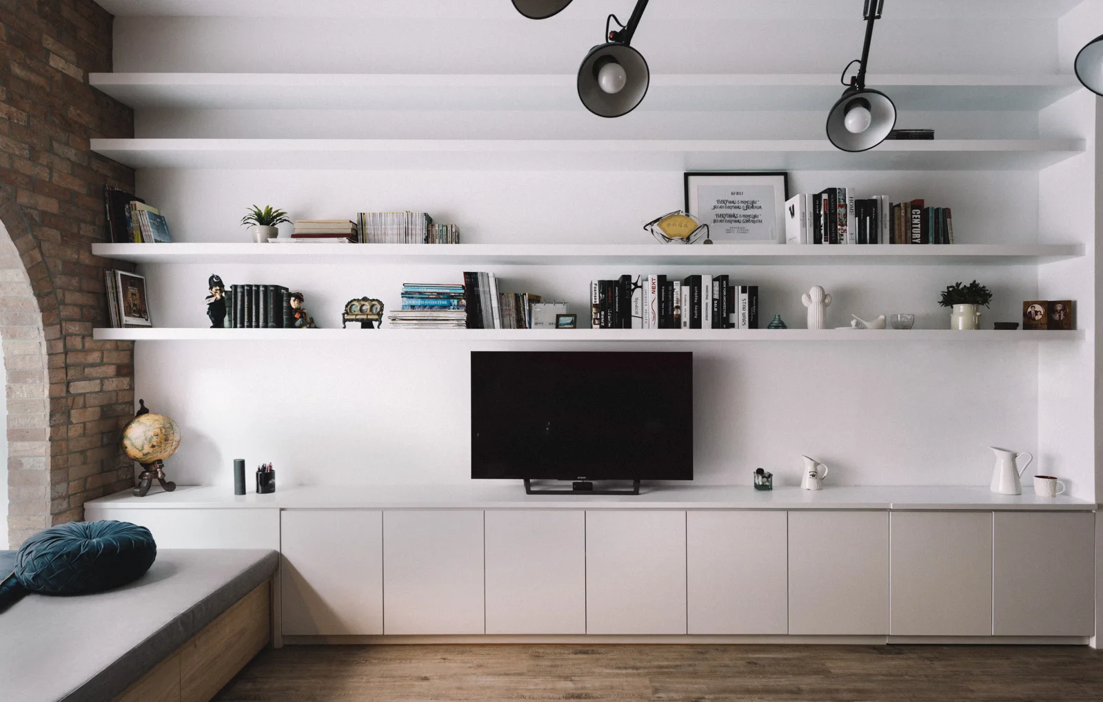 A white built-in media unit with floating shelves and flat-panel base cabinets stands beside an exposed brick archway.