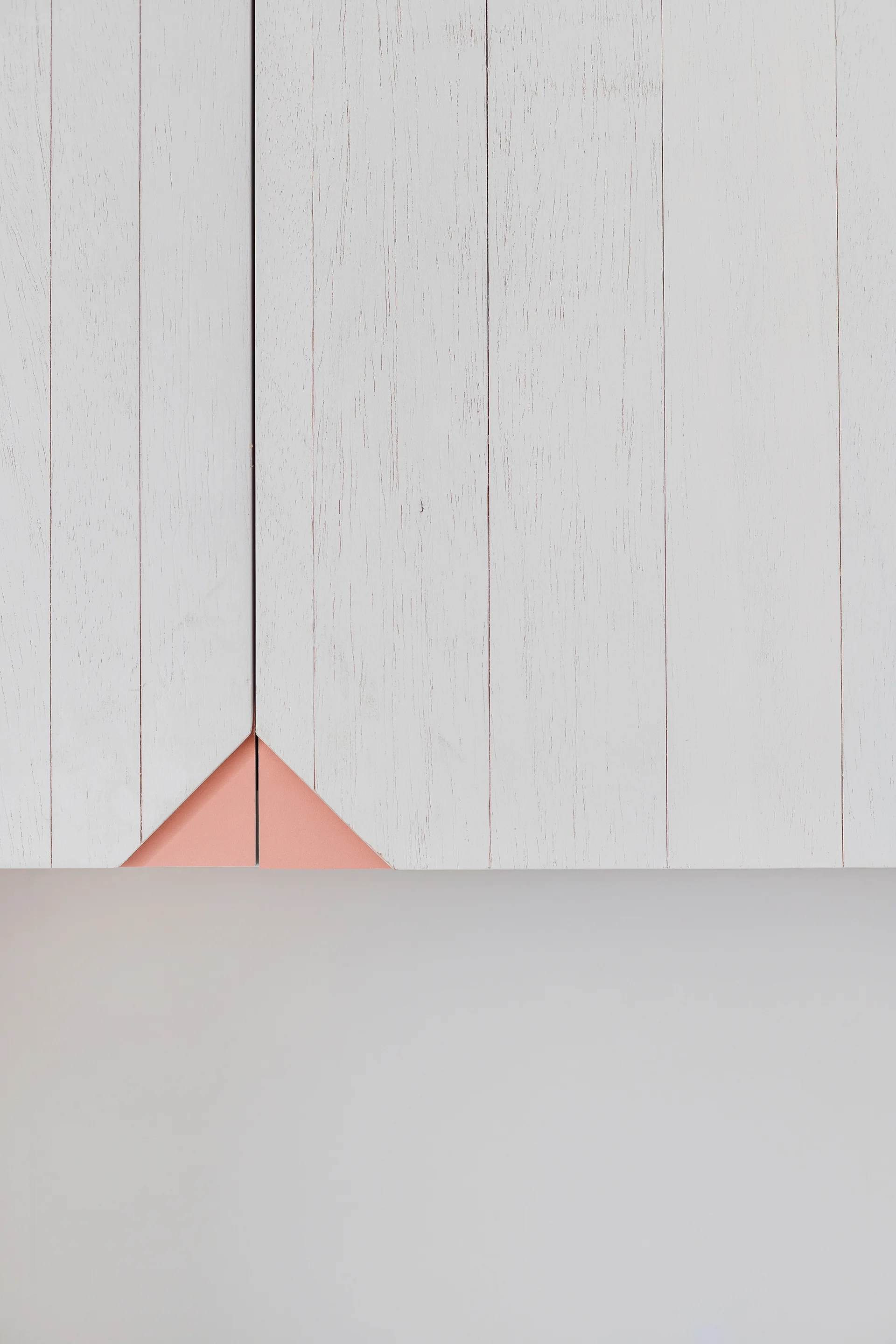 A close-up of off-white vertical wood paneling featuring a small recessed triangular cutout with a matte coral pink interior.