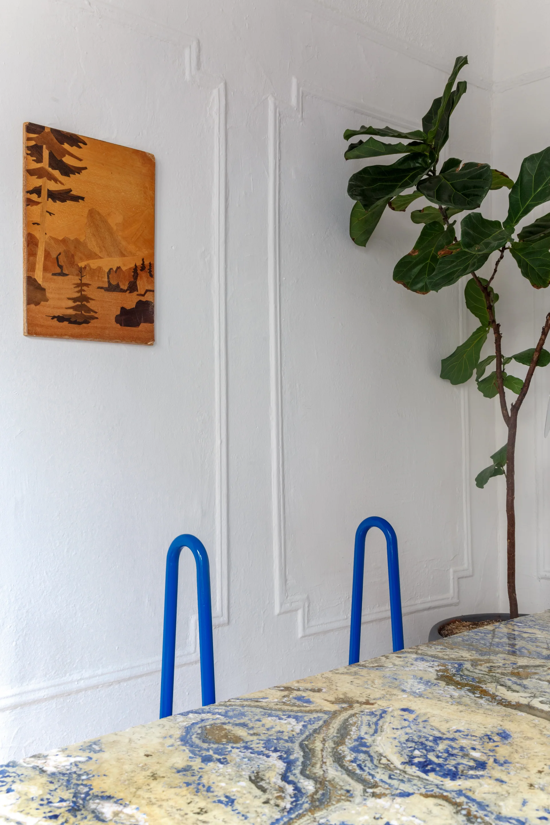 Cobalt blue tubular metal chairs sit behind a patterned blue and yellow stone table in a room with white paneled walls.