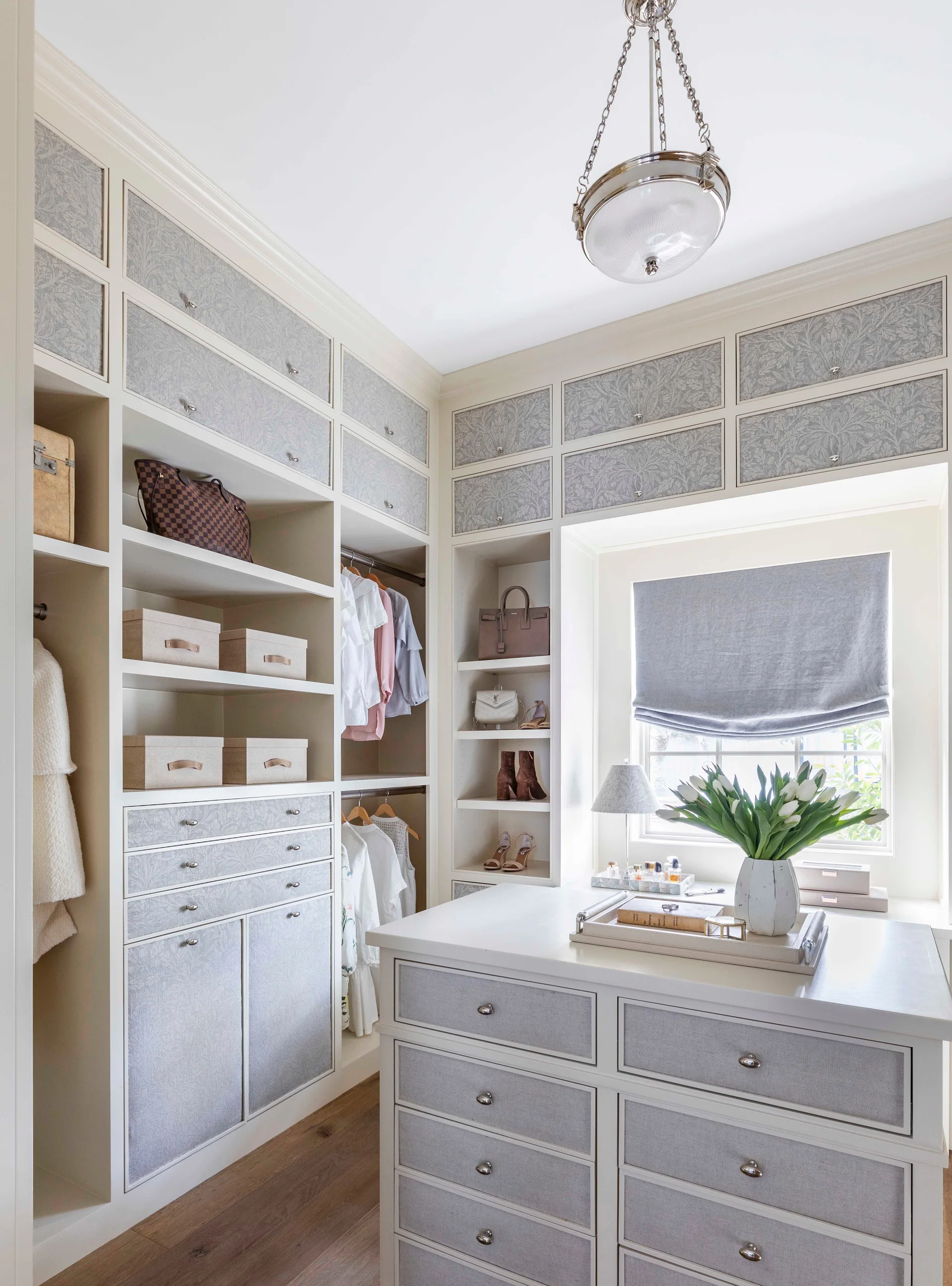 A bright walk-in closet features built-in off-white cabinetry with floral patterned grey drawers, a central island, and a window with a roman shade.