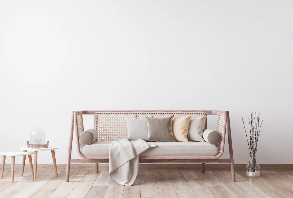 A minimalist light wood and rattan sofa sits against a plain white wall next to small white nesting tables and a vase of branches.