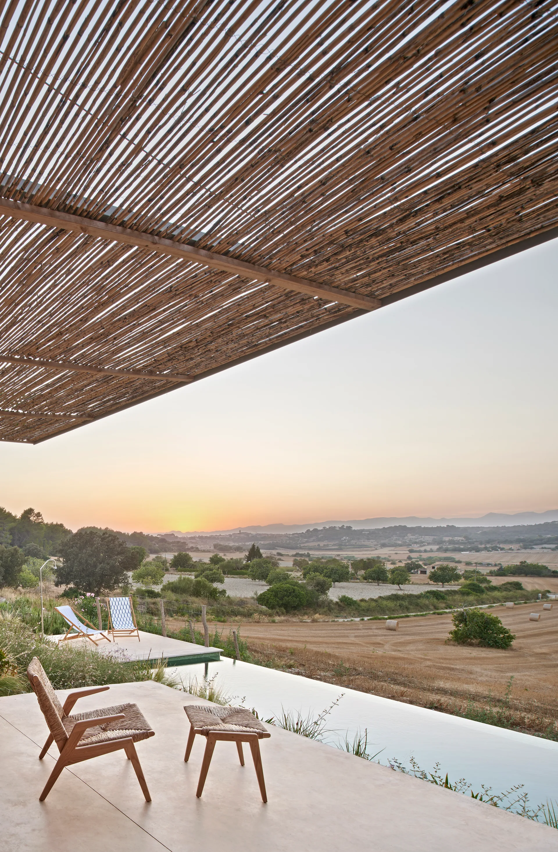 A concrete patio features a woven wooden armchair, matching ottoman, and a slatted cane pergola overlooking an infinity pool and rural sunset landscape.