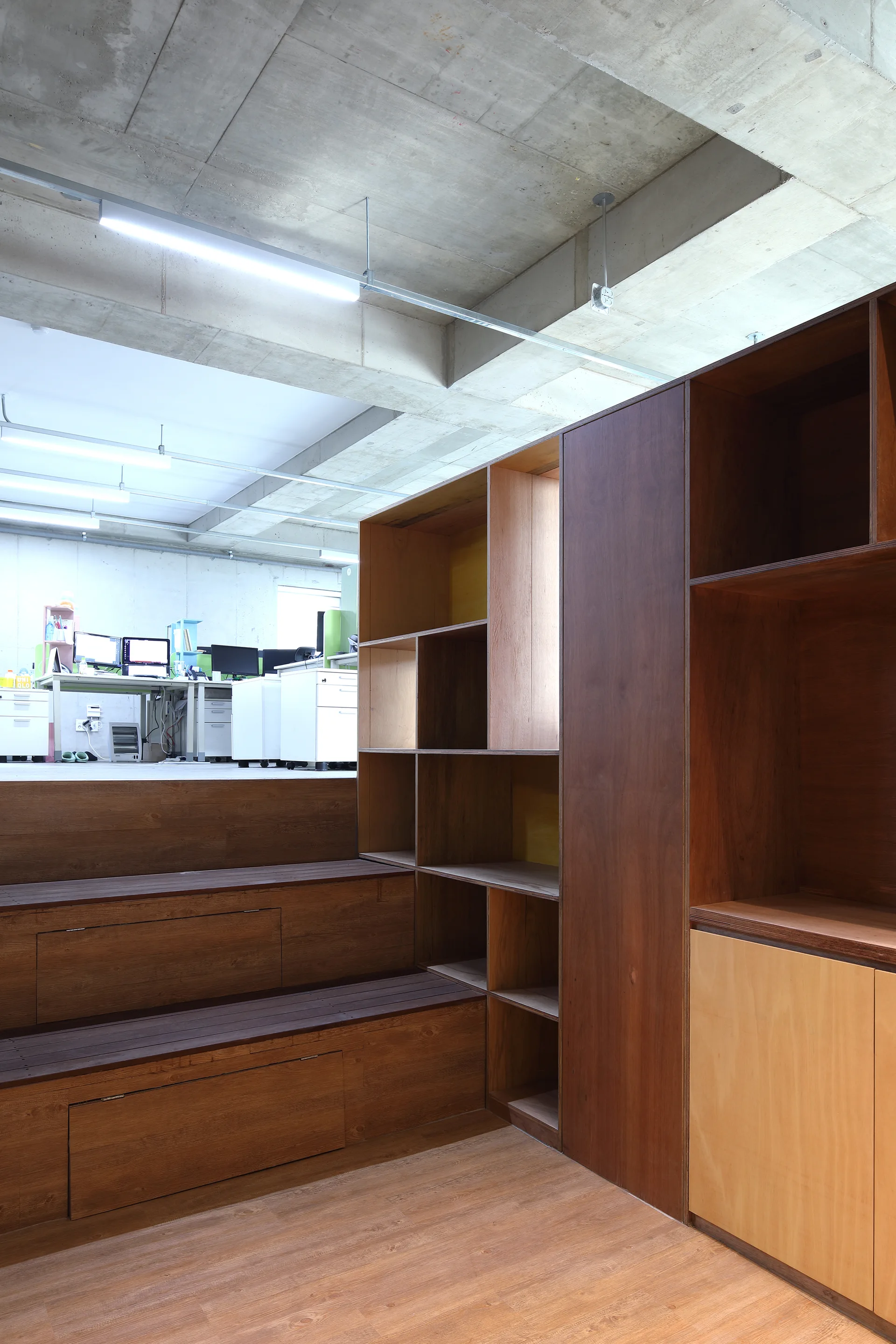 A modern office space features a large wooden custom shelving unit, tiered plywood platform seating, and an exposed concrete ceiling with linear lights.