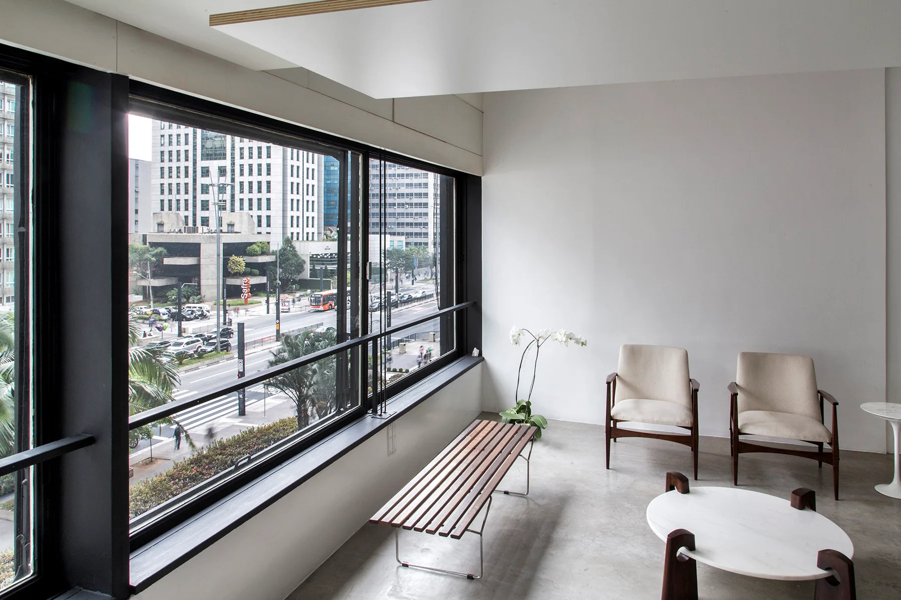 Two cream armchairs and a slatted wooden bench stand near a large window overlooking a city street.