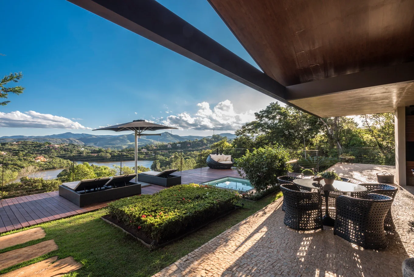 A modern outdoor terrace features a wooden deck with lounge chairs and a stone-paved dining area overlooking a green valley and lake.