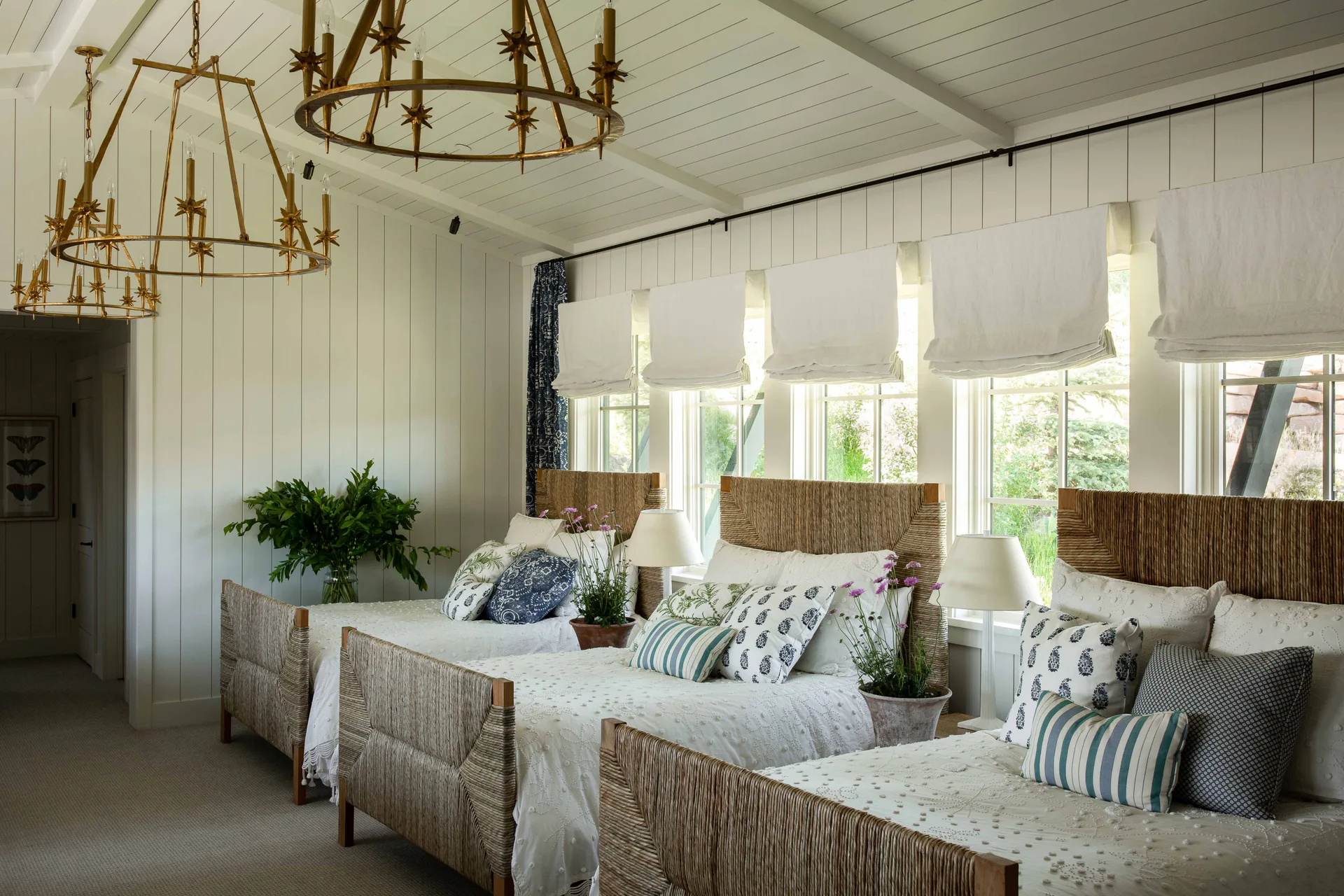 Three twin beds with woven headboards sit side-by-side in a bedroom with white paneled walls, brass star-shaped chandeliers, and windows.