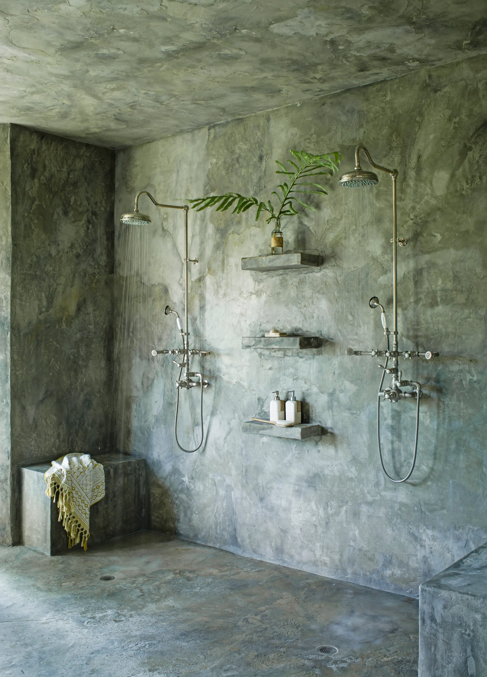 A walk-in shower with textured concrete walls, ceiling, and floor features two running chrome shower heads and three integrated floating shelves.