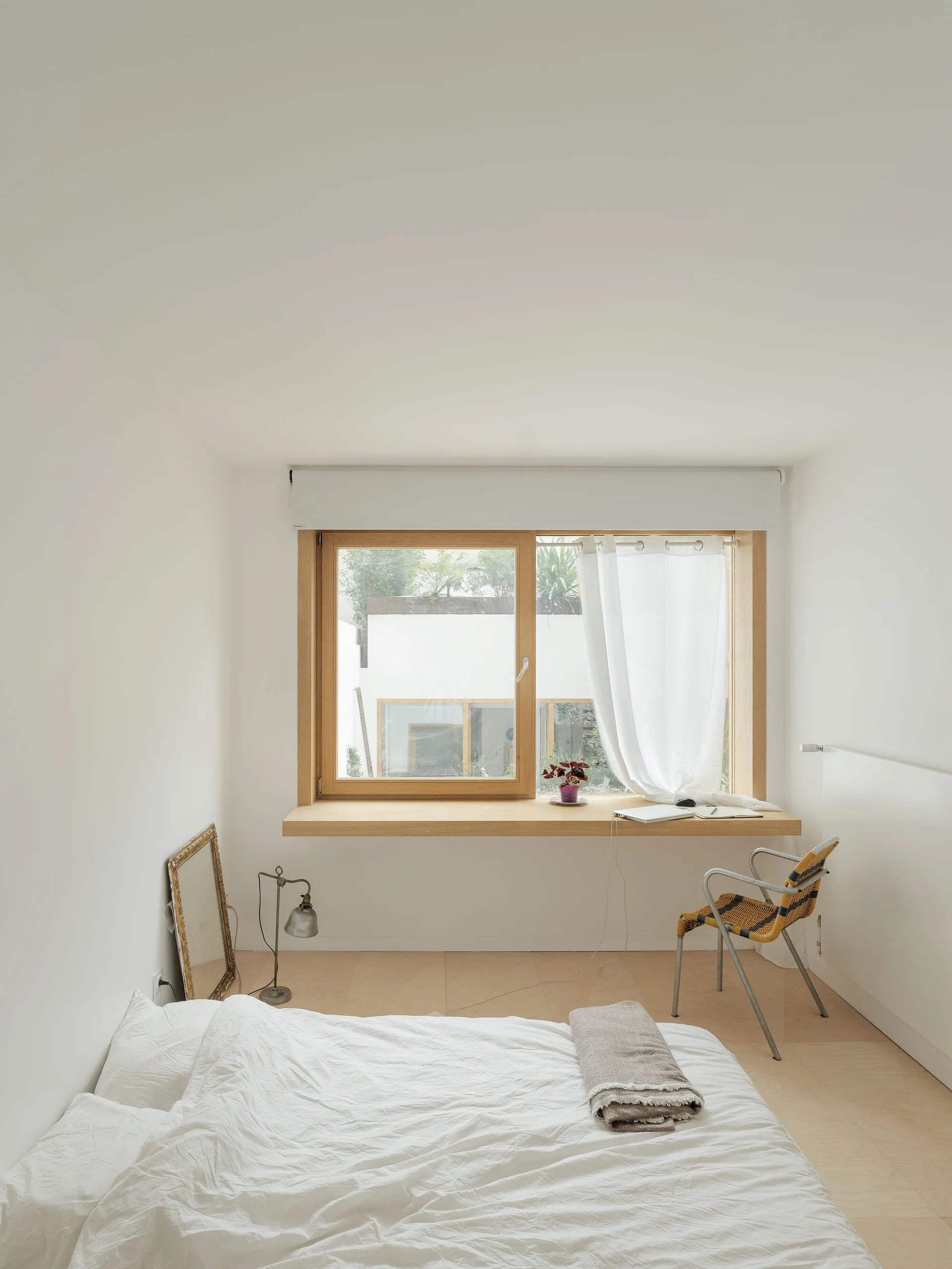 A minimalist bedroom features a low bed with white linens, a woven chair, and an extended wooden window sill serving as a desk.