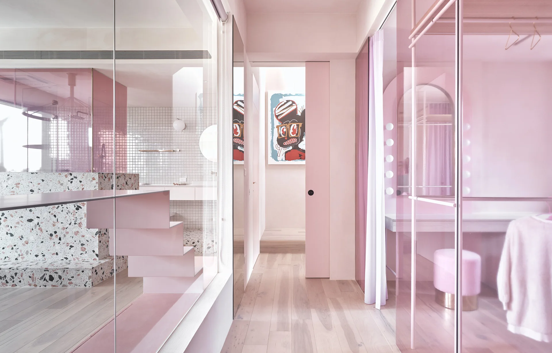 A pink dressing area featuring pink tinted glass wardrobe doors, a terrazzo platform, a vanity mirror with globe lights, and light wood flooring.