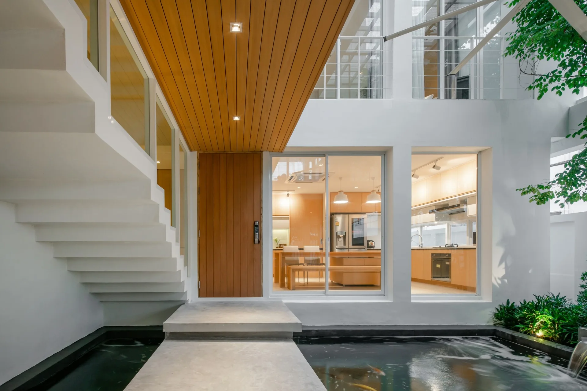 A modern home entryway features a concrete stepping path over a koi pond, next to a white staircase and a tall wooden front door.