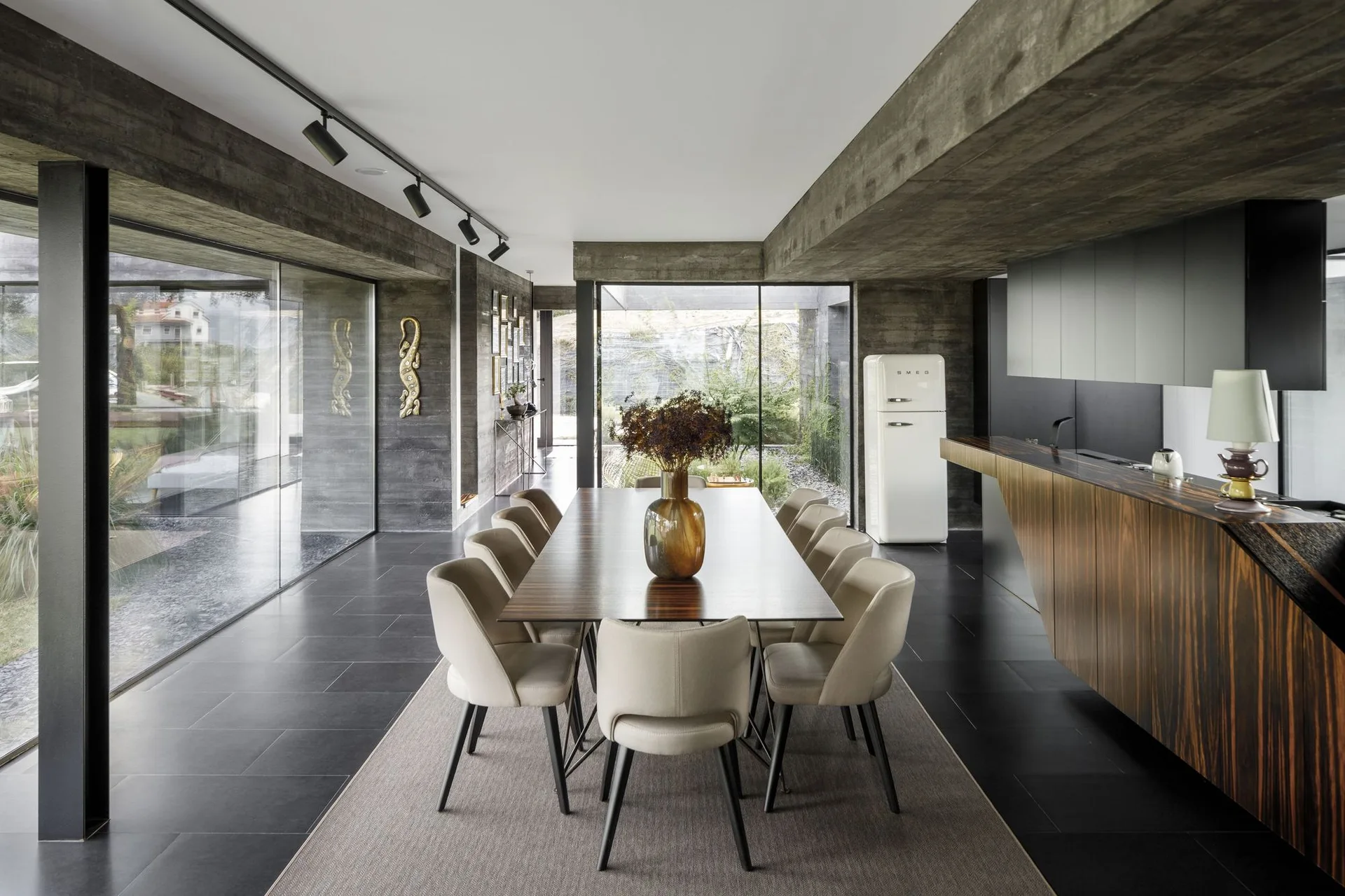 A modern dining room features a long dark wood table, beige upholstered chairs, and raw concrete walls next to large glass windows.