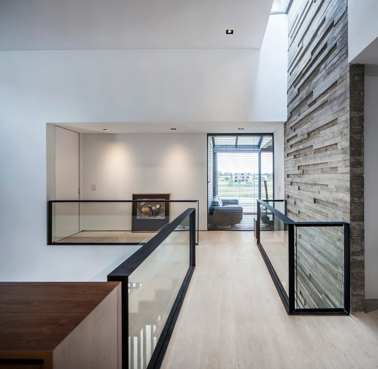 A modern home landing features a textured concrete accent wall, glass panel railings with black metal frames, and light stone flooring under a skylight.