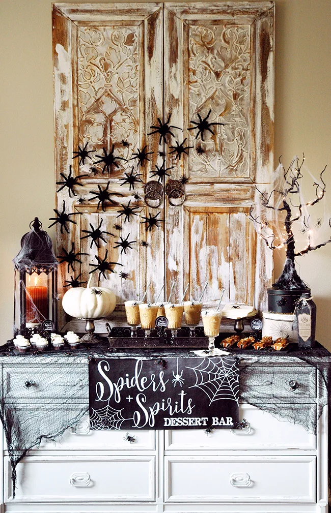 A Halloween dessert bar is arranged on a white painted dresser in front of a distressed, carved wooden backdrop decorated with faux spiders.