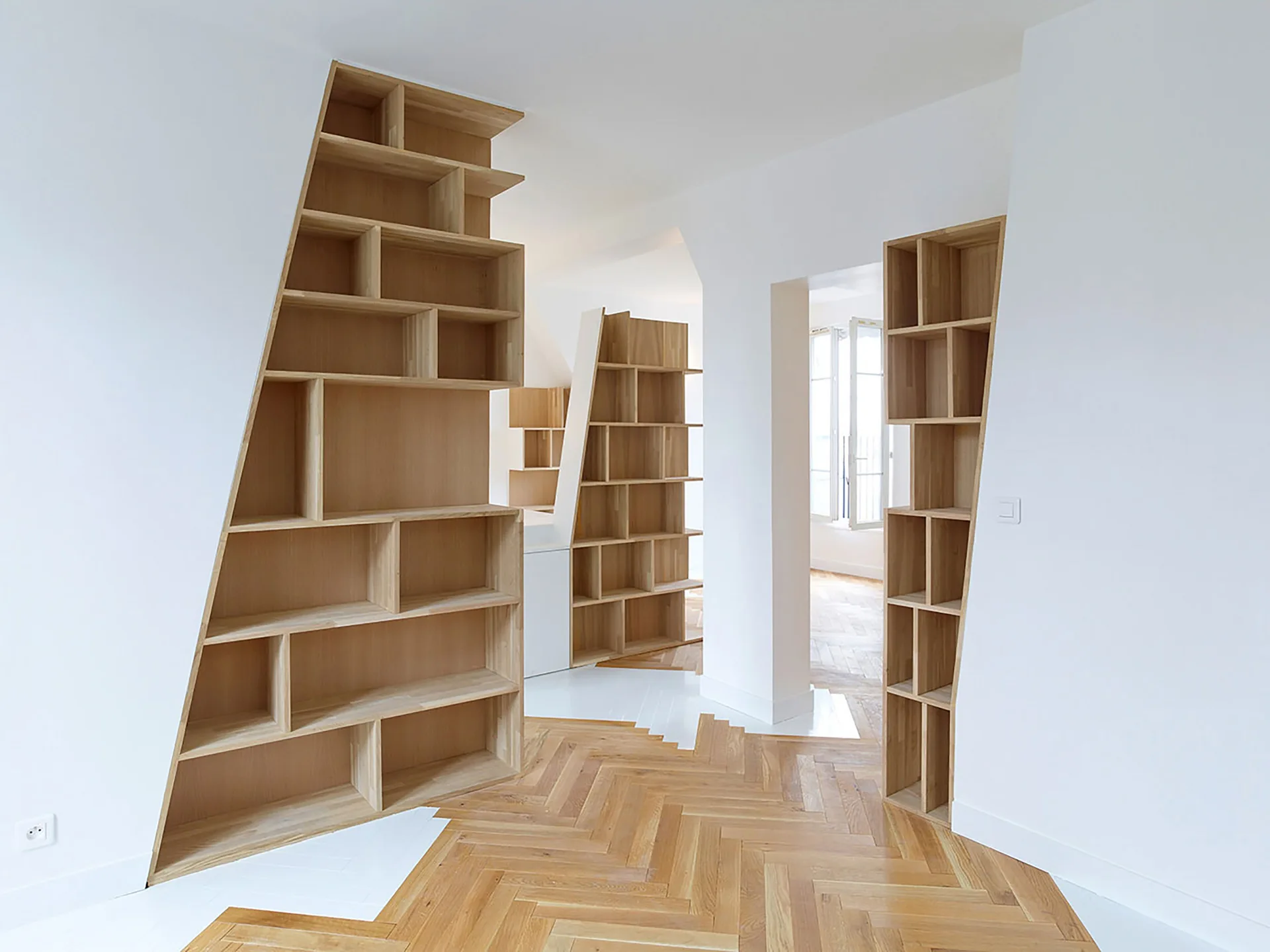 Custom light wood bookshelves with slanted sides are integrated into white hallway walls above a herringbone pattern parquet floor.