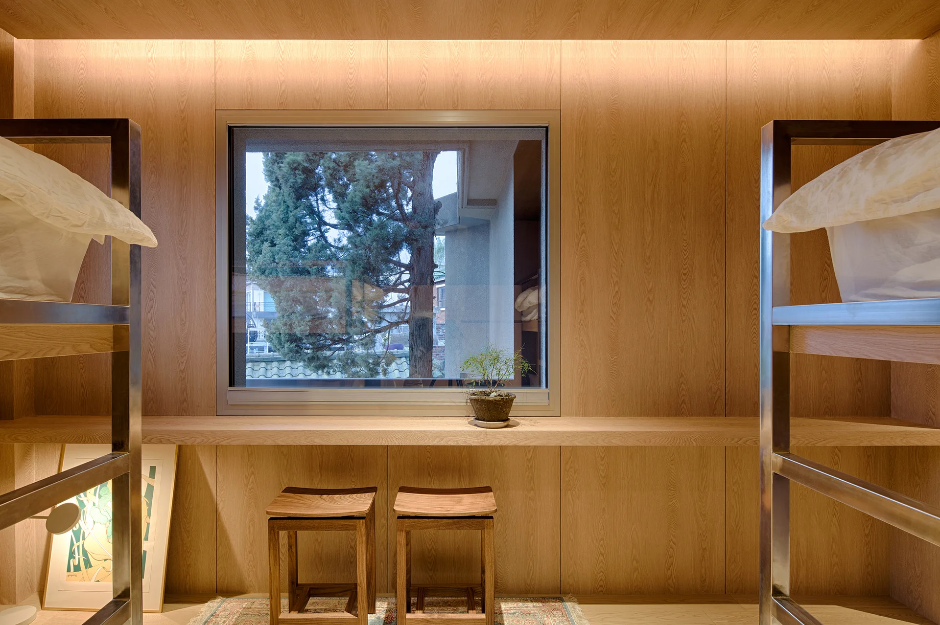 A minimalist wood-paneled bedroom features a large window with a built-in ledge, two wooden stools, and integrated warm lighting.