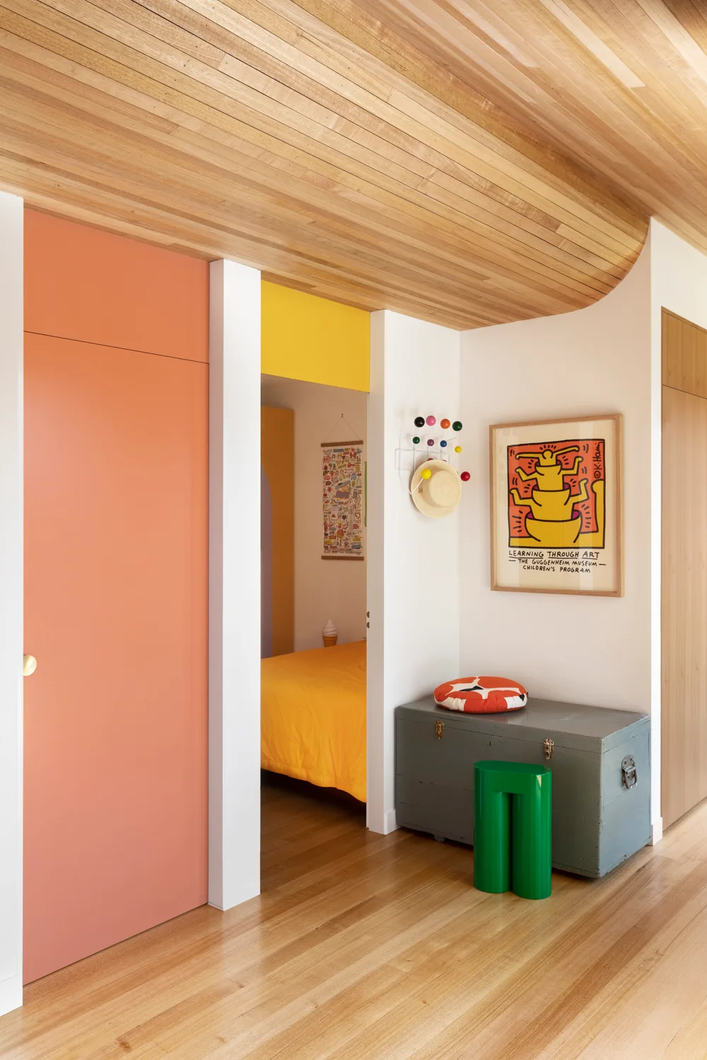 A hallway with a curved wood plank ceiling features a bright orange door, a gray storage chest with a green stool, and a doorway leading to a yellow bedroom.