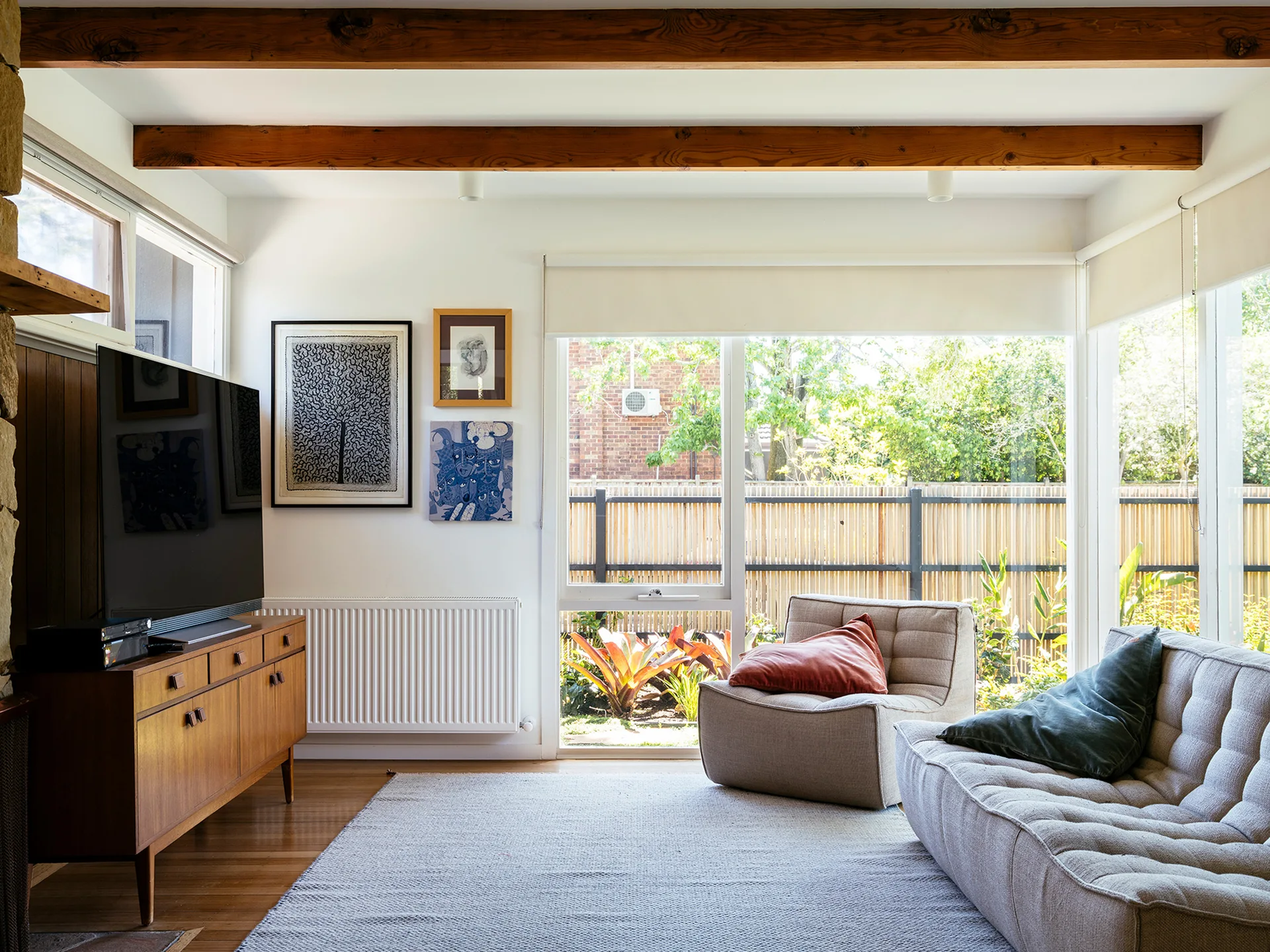 A bright living room features mid-century modern furniture, exposed wooden ceiling beams, and large windows looking out to a garden.