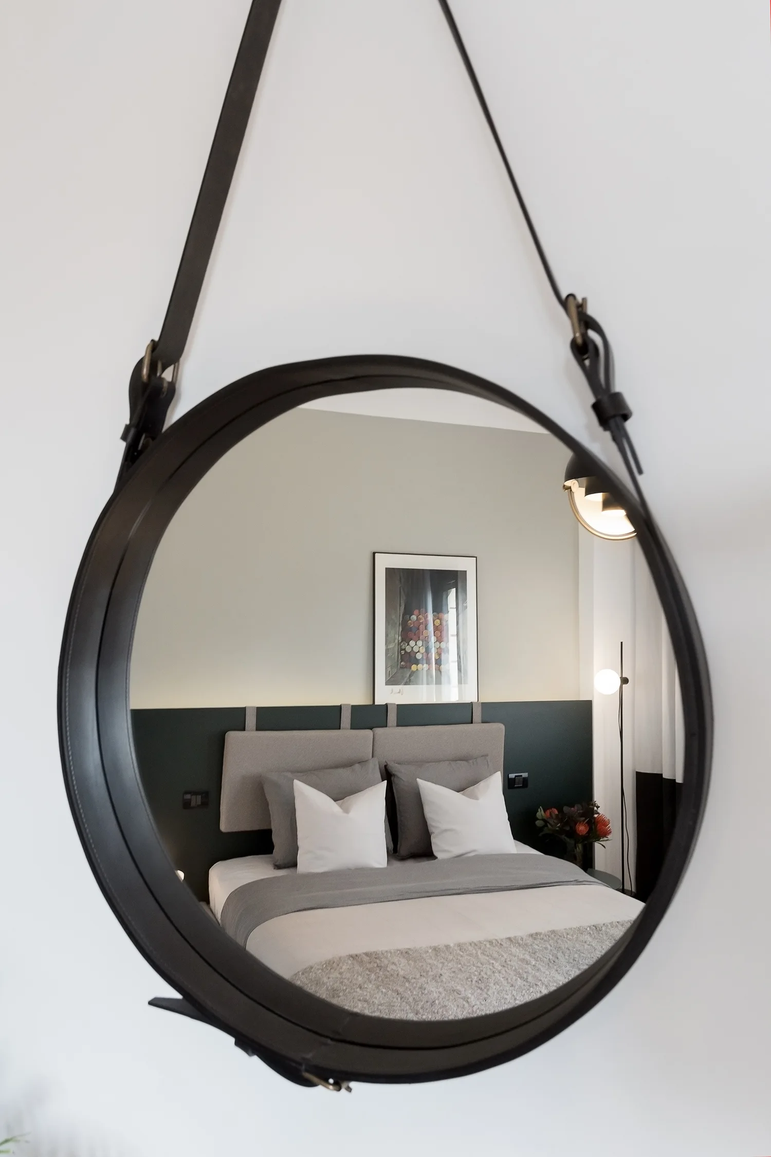 A large circular mirror with a black leather strap hangs on a white wall, reflecting a grey and white bed set against a dark green accent wall.