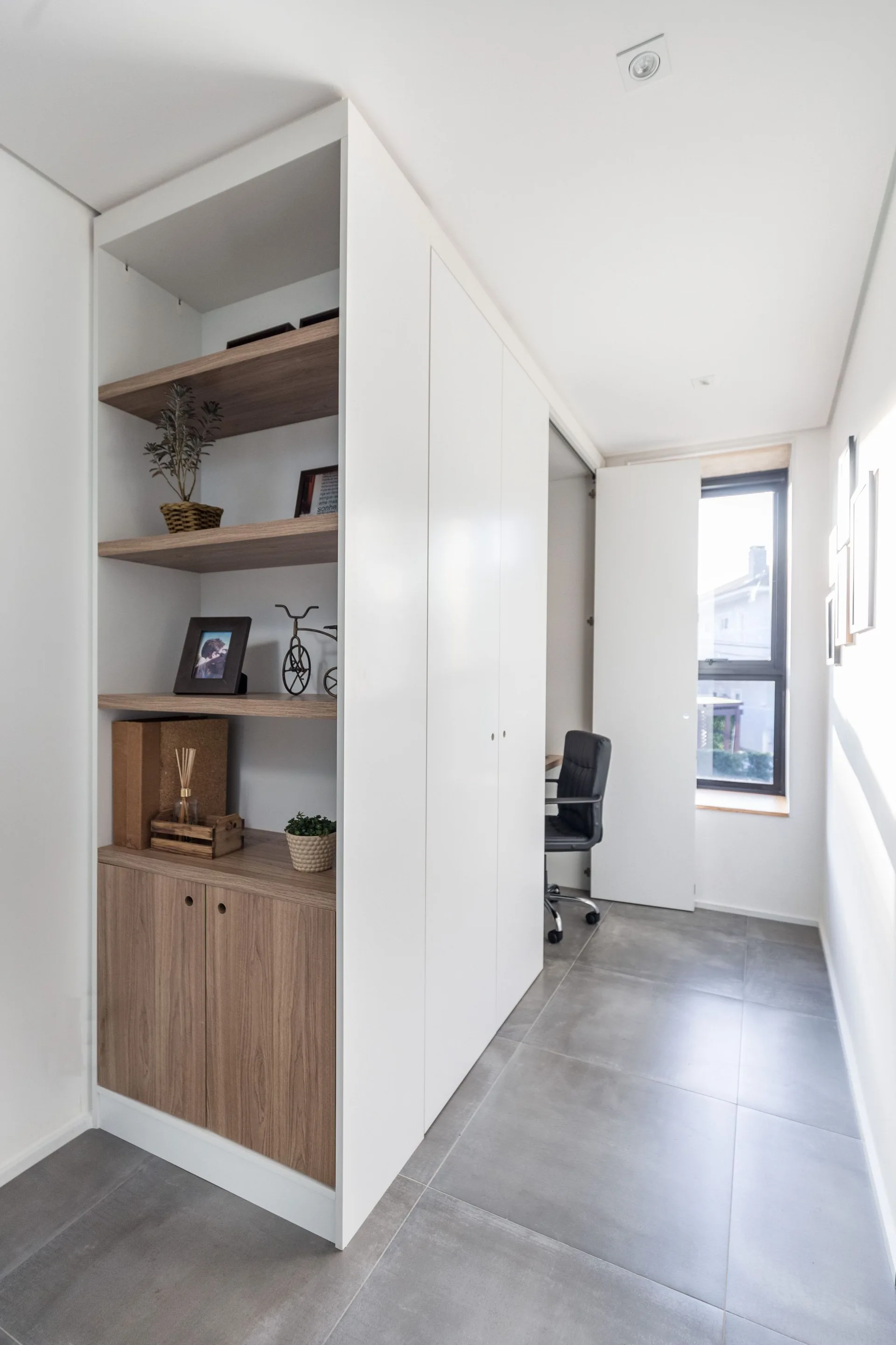 A tall white storage cabinet features open wooden shelves styled with decor next to a home office nook with a black chair and gray tile flooring.