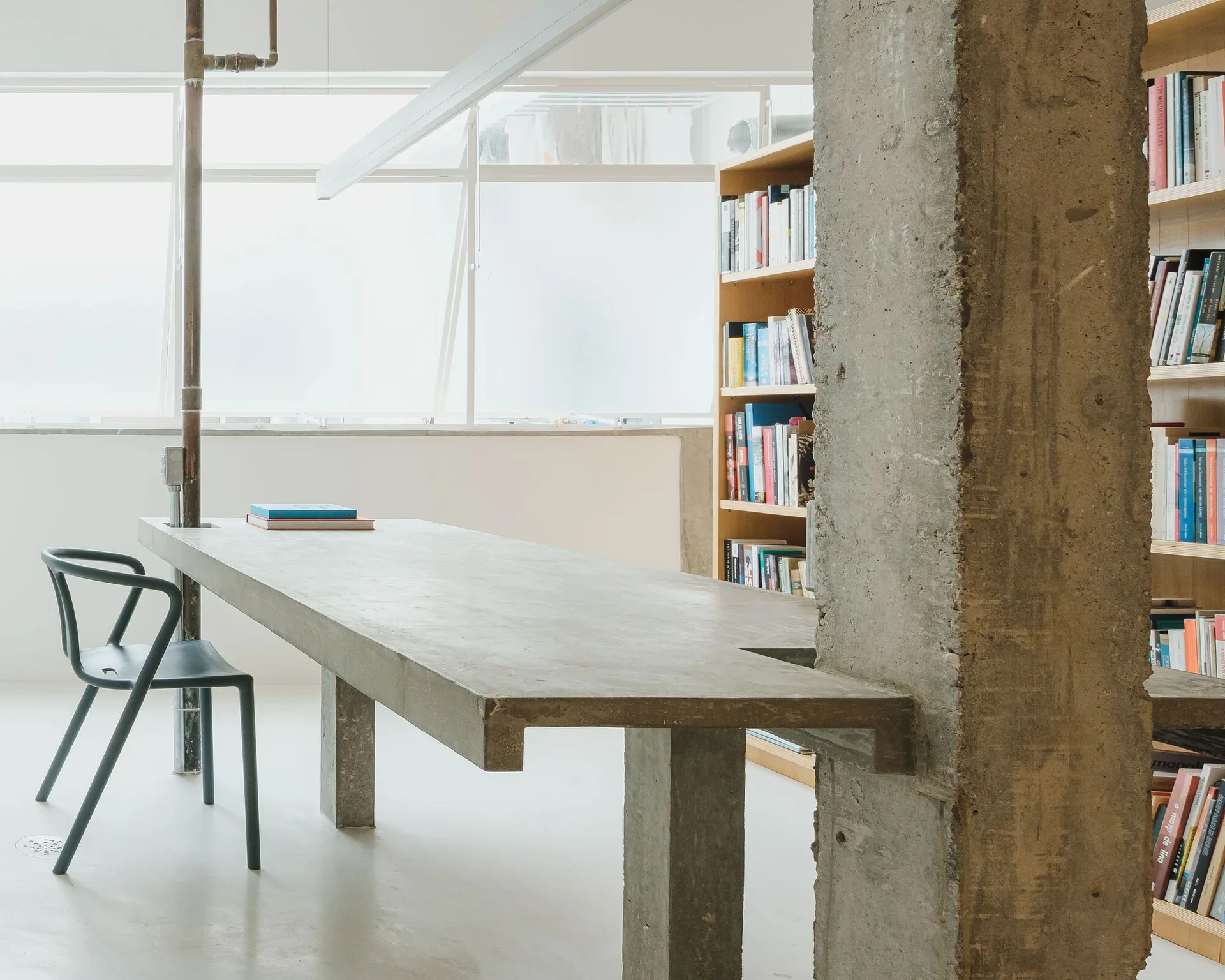 A long concrete desk stands next to a raw concrete column and a black modern chair in a minimalist, naturally lit workspace.