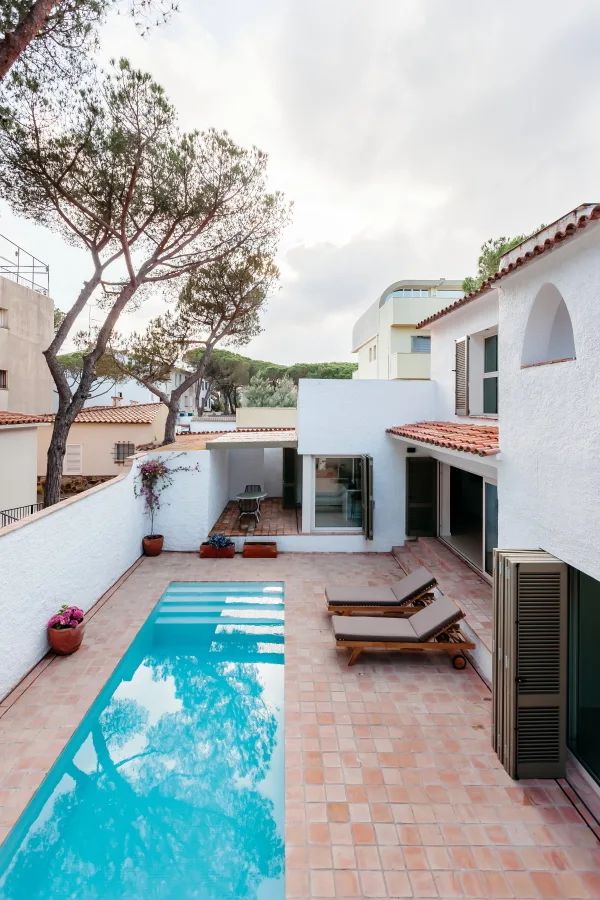 A rectangular swimming pool is set in a terracotta tiled courtyard next to a white stucco house with louvered shutters and wooden lounge chairs.