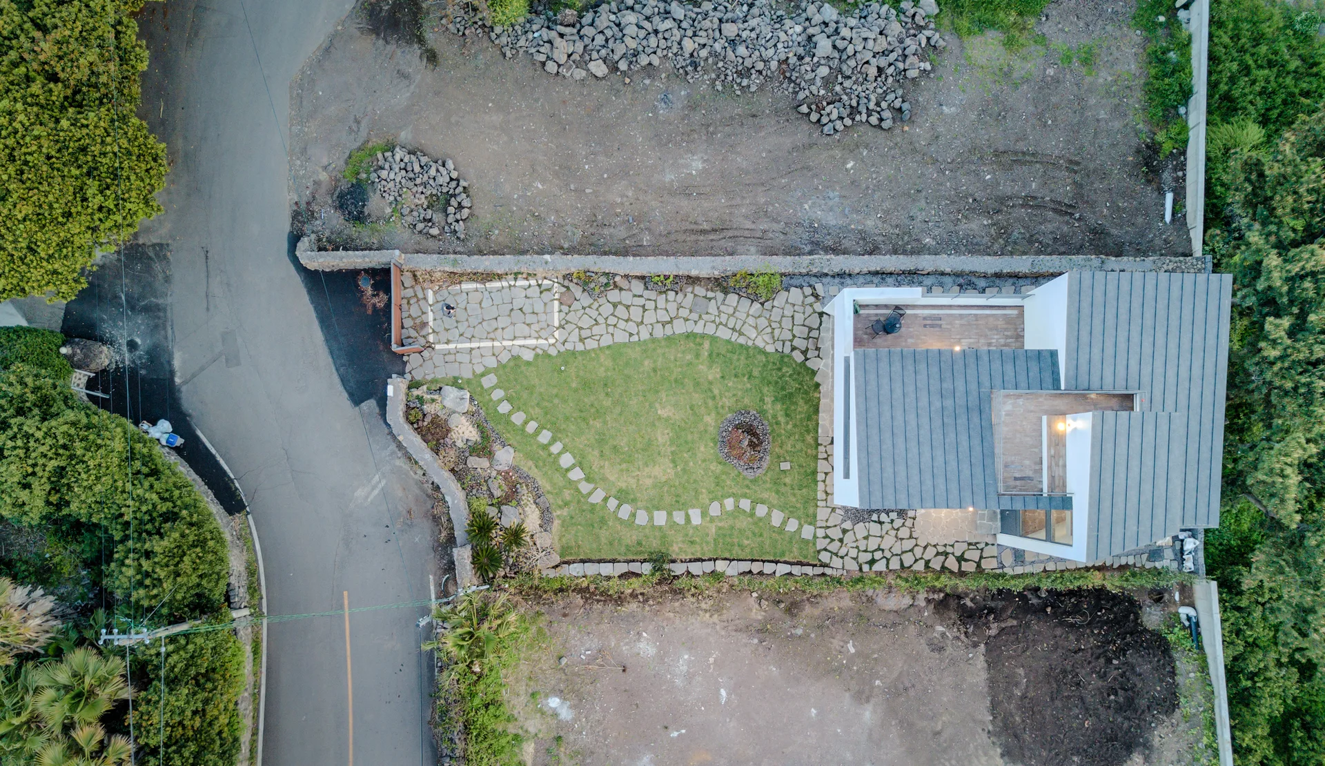 An overhead aerial view of a modern residence featuring a multi-level gray metal roof, a curved green lawn, and winding stone walkways.