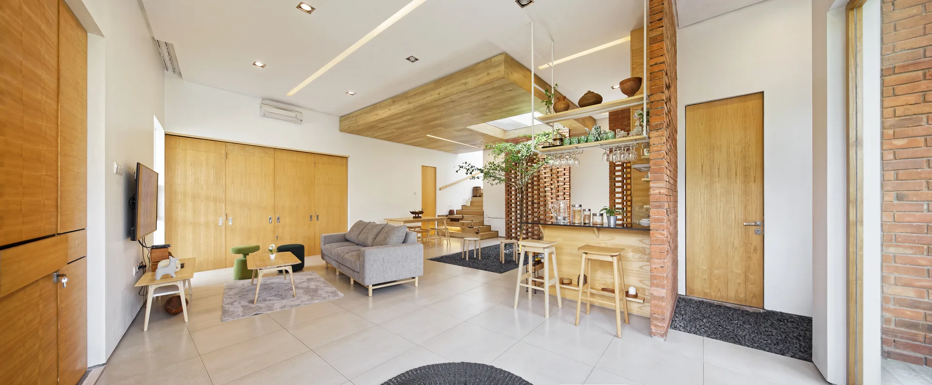 A spacious open-concept living room features a grey sofa, a wooden bar with stools against an exposed brick wall, and an indoor tree in a central lightwell.