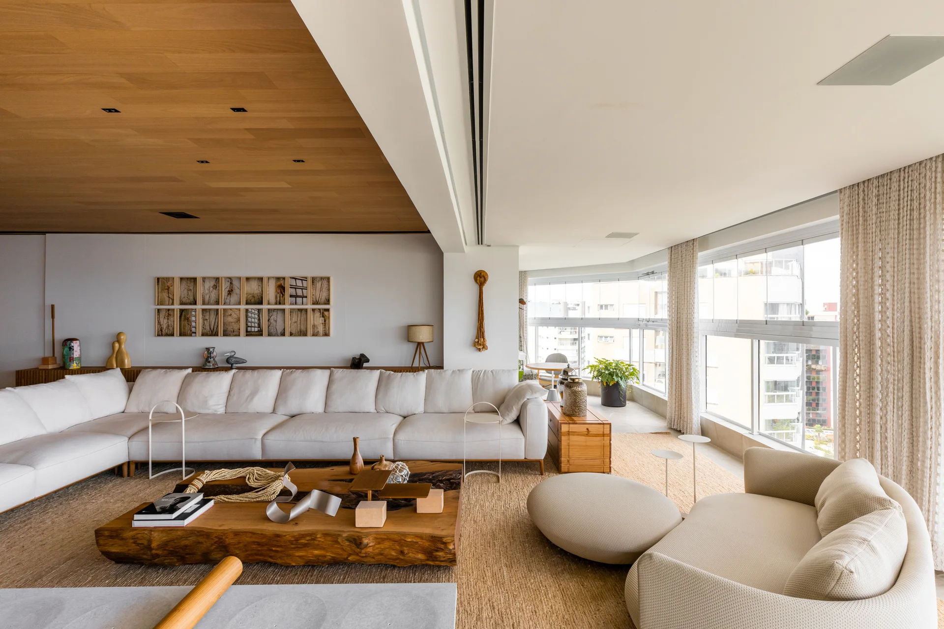 A bright living room features a long cream sectional sofa, a large live-edge wood coffee table, a woven rug, and a wood paneled ceiling.