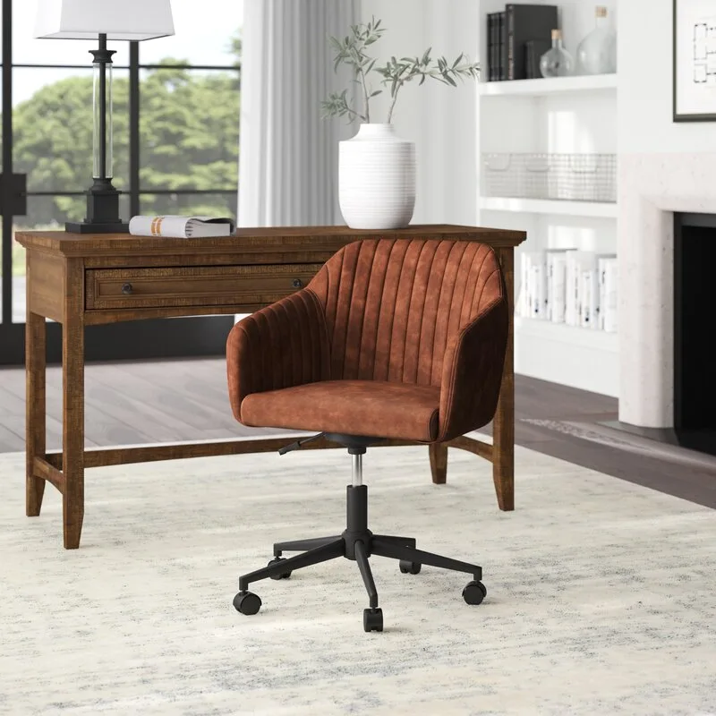 A rust-colored channel-tufted desk chair on wheels sits in front of a wooden writing desk on a light distressed area rug.