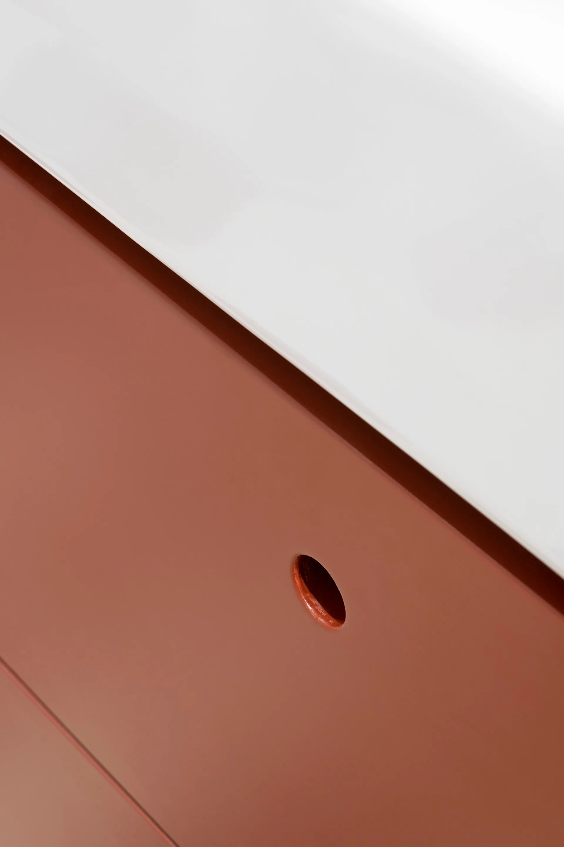 A close-up of minimalist two-tone cabinetry featuring a white upper surface and a matte terra-cotta drawer front with a circular recessed pull.