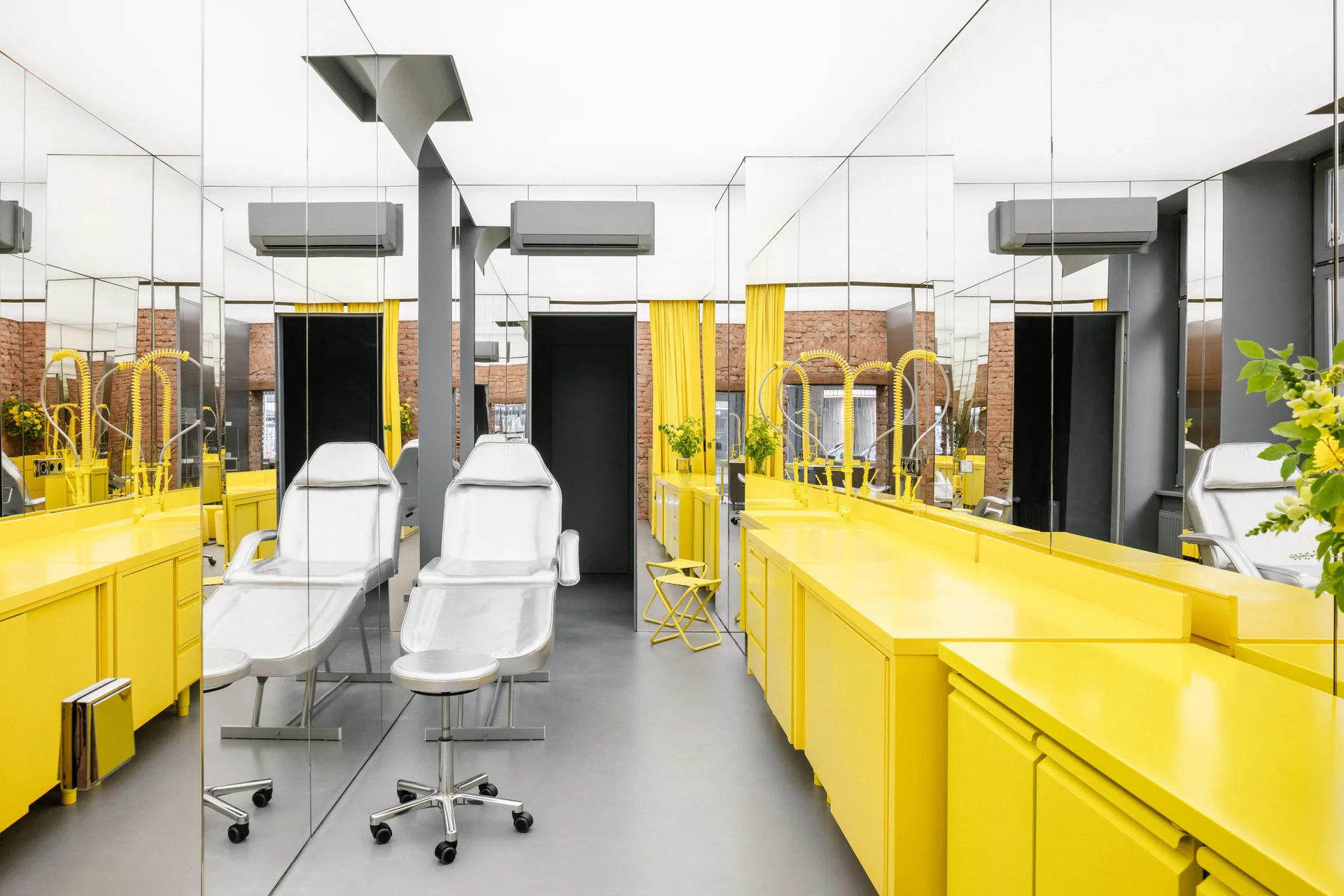 A minimalist treatment room featuring bright yellow cabinetry, two white medical chairs, and expansive mirrored walls reflecting the entire space.