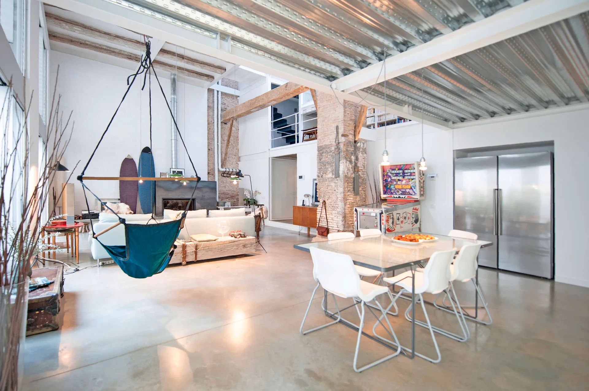 An open industrial loft features a dining area with a glass table, a concrete floor, a hanging fabric swing, and exposed brick columns.