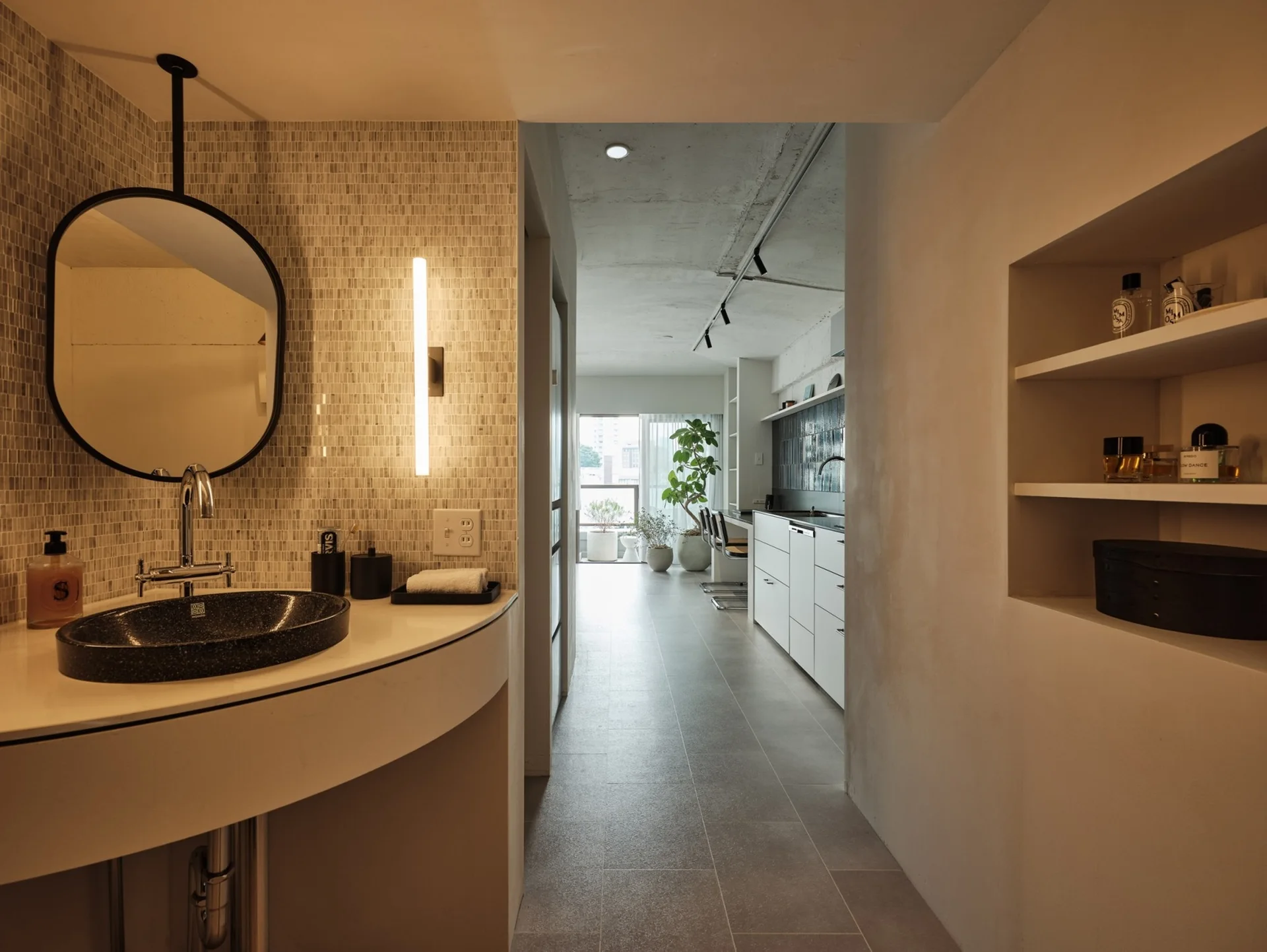 A curved bathroom vanity with a black sink and round mirror stands in a hallway that leads toward a bright kitchen.