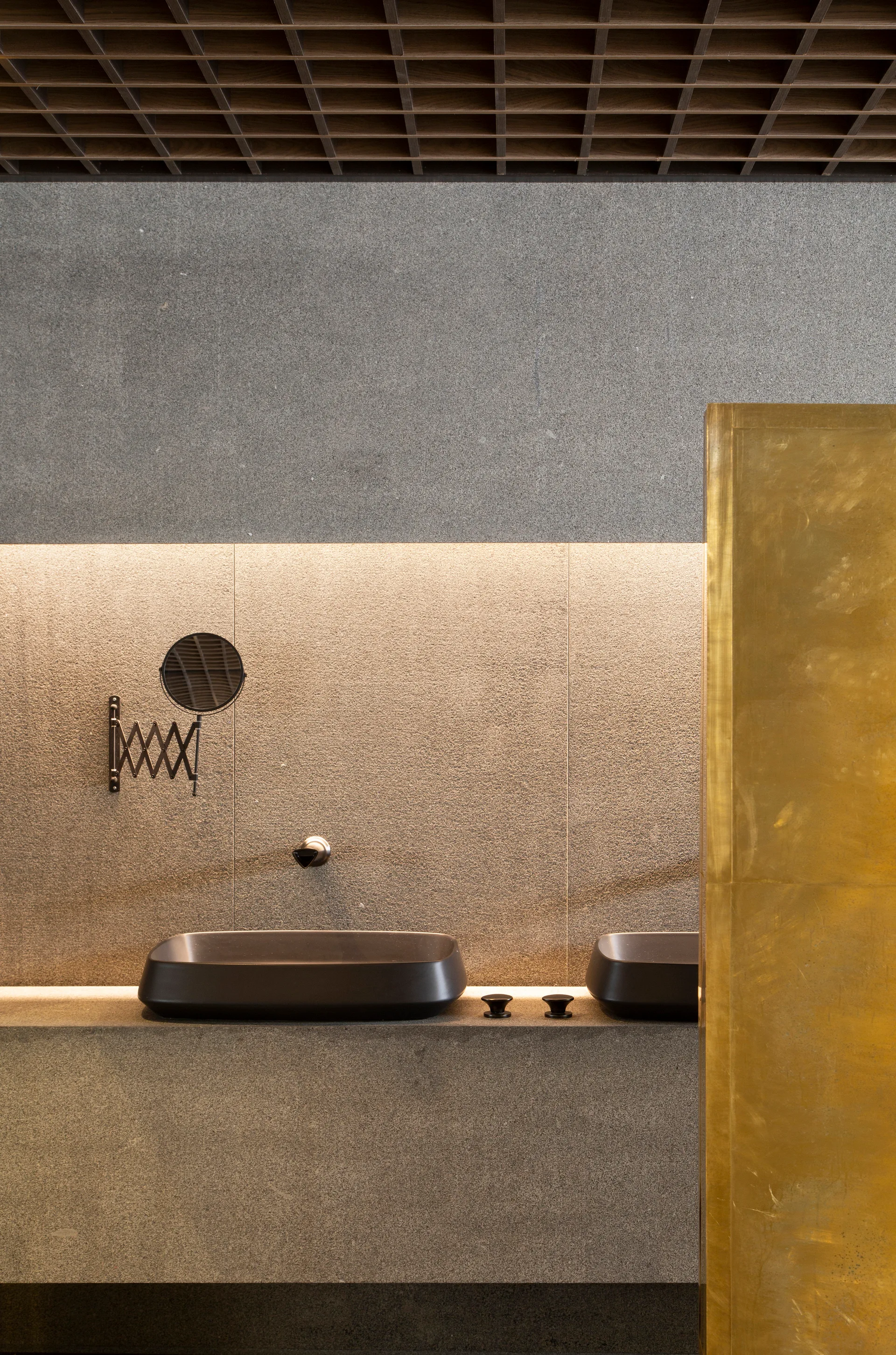 A modern grey stone bathroom featuring two black vessel sinks, a gold partition, and a dark wooden grid ceiling.