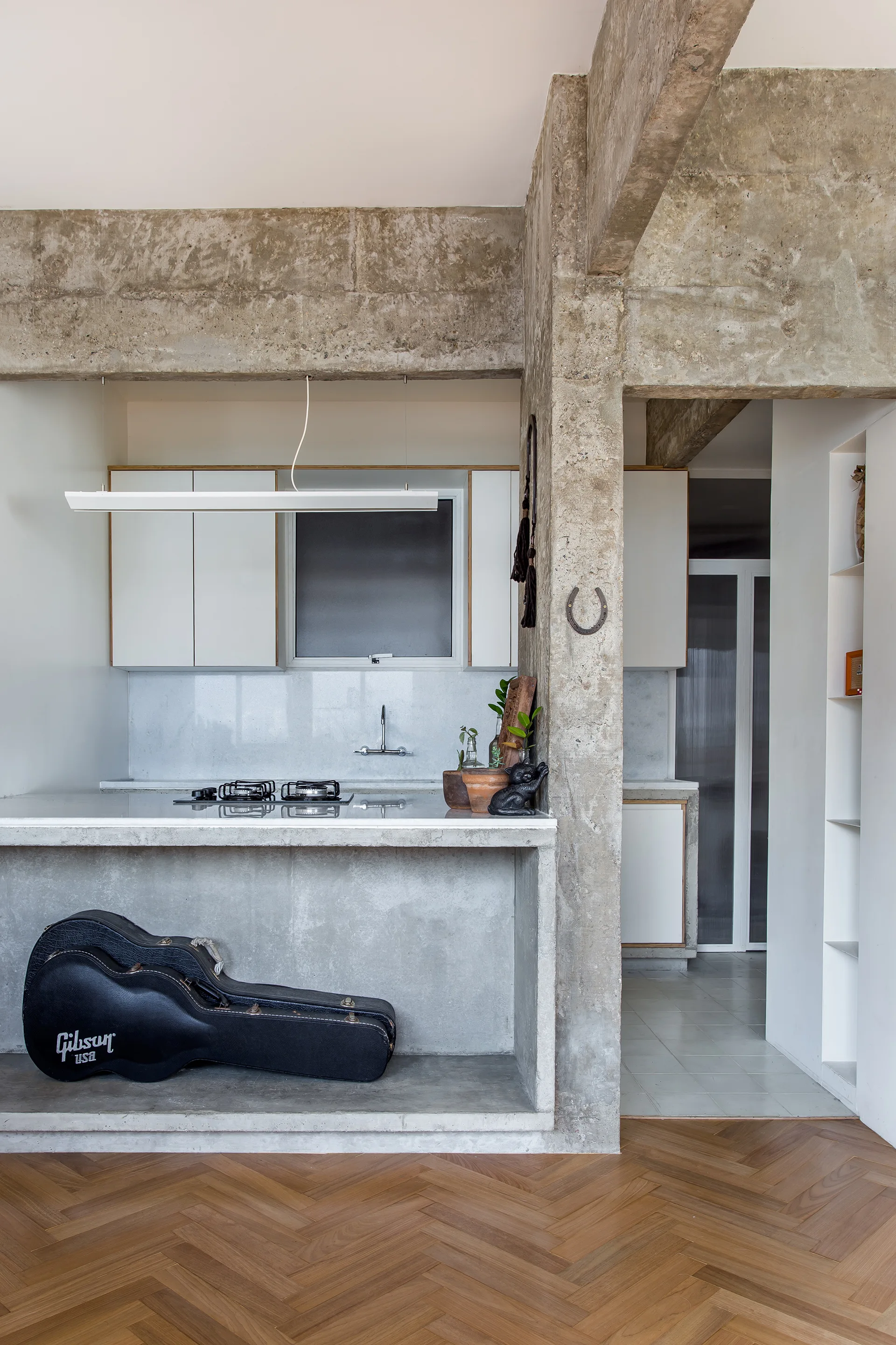 A modern minimalist kitchen with raw concrete pillars, white cabinets, a concrete island, and warm wood herringbone floors.