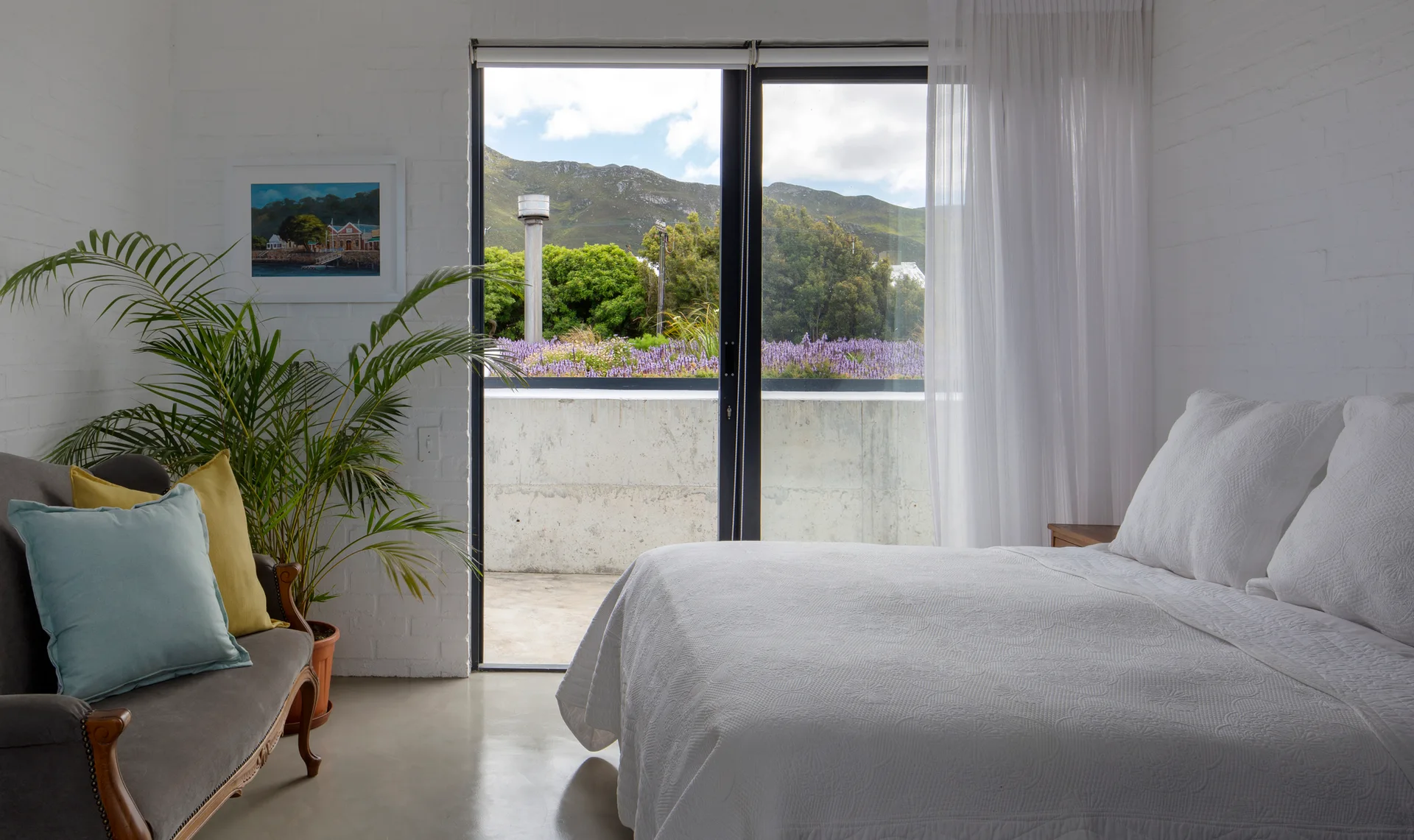 A minimalist bedroom features a white bed, a grey vintage settee, and sliding glass doors opening to a concrete balcony with mountain views.