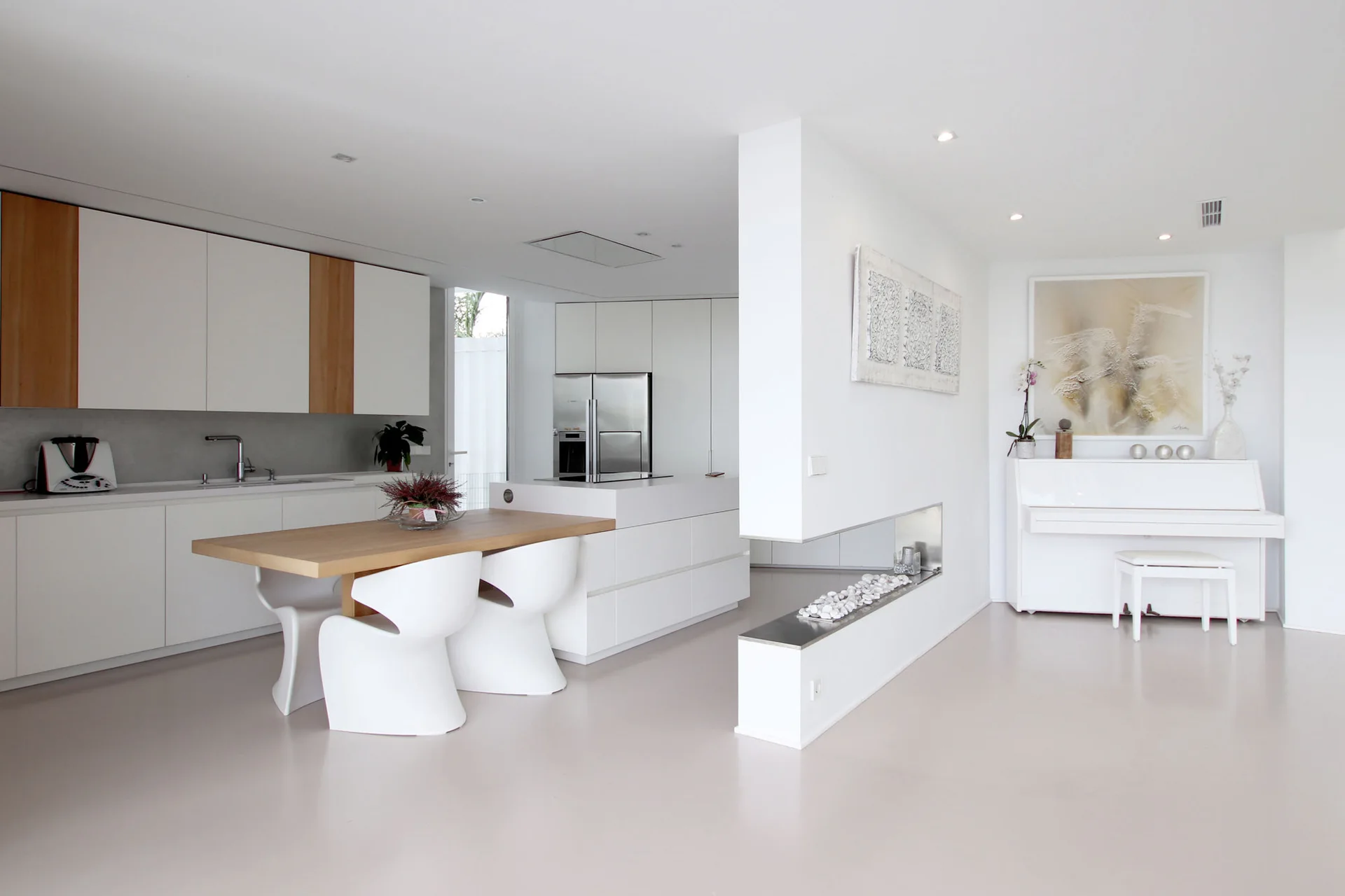 A minimalist white open-plan kitchen features a light wood dining table with curved chairs, a low room divider, and a white upright piano.