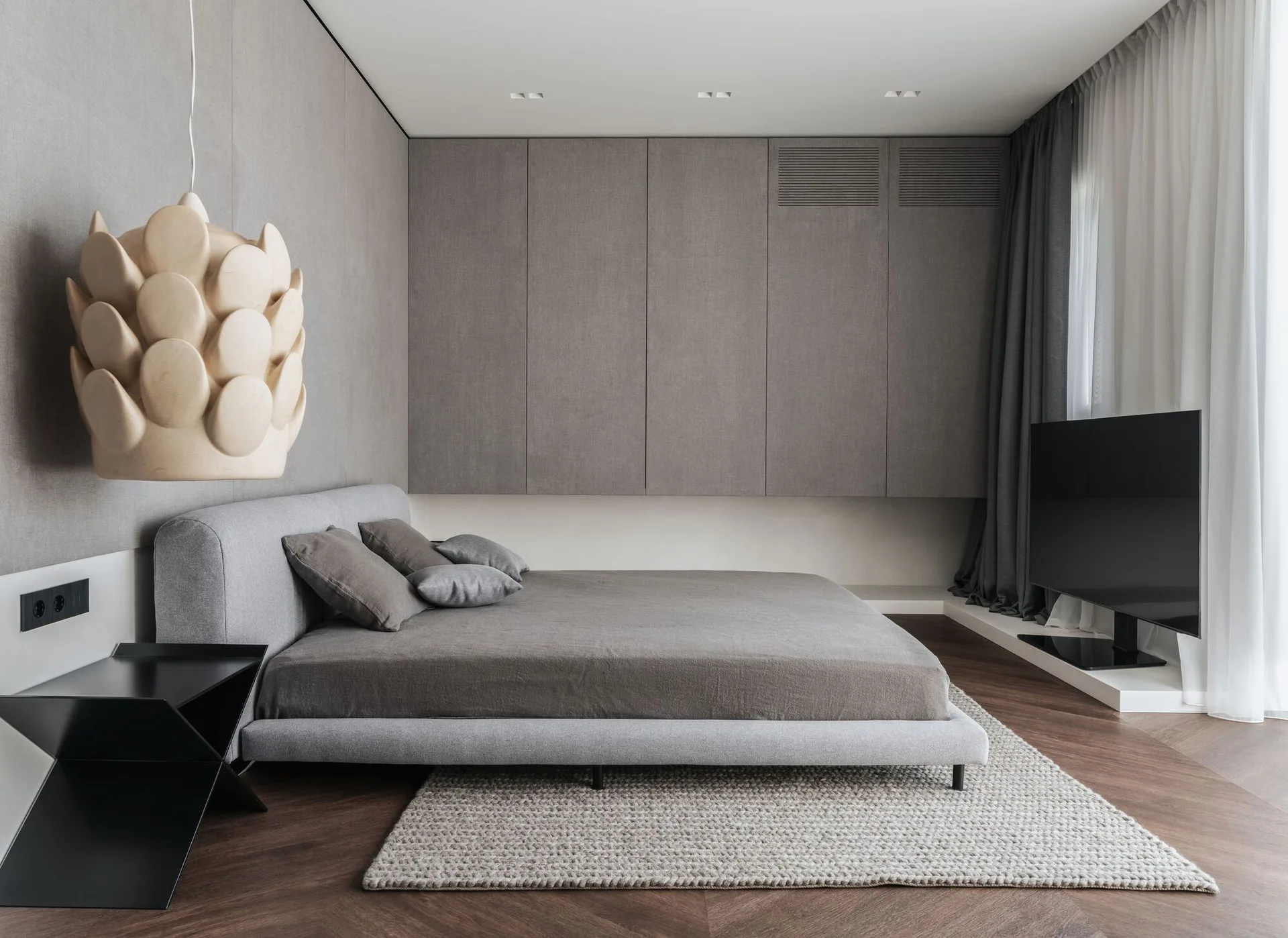 A modern bedroom features a grey upholstered platform bed, fabric-wrapped wall panels, integrated storage cabinets, and a black sculptural pendant light.