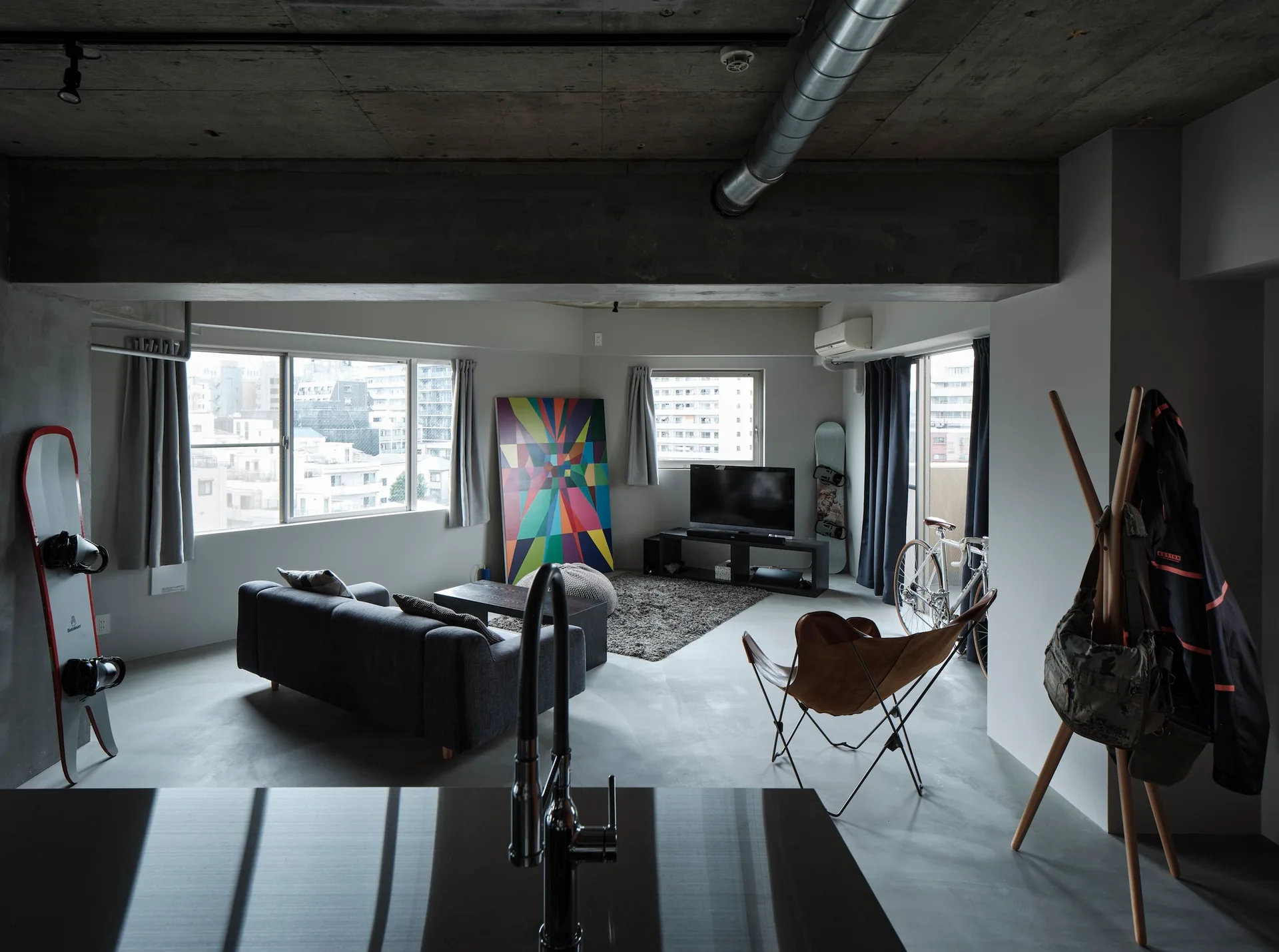 An industrial apartment living room features a dark sofa, a leather butterfly chair, and an exposed raw concrete ceiling.