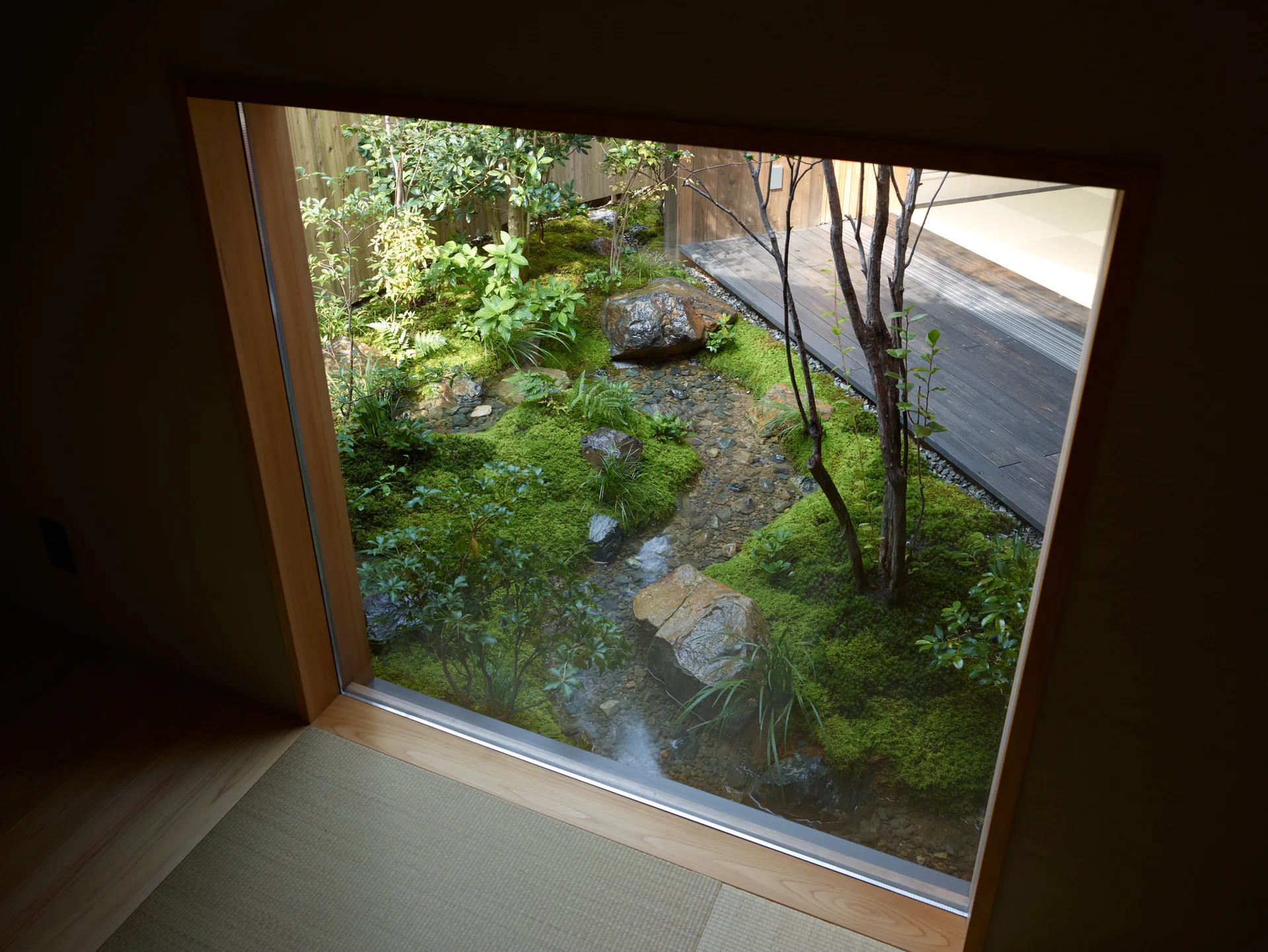 A view through a large wood-framed window looking out at a lush courtyard moss garden with a small pebble stream, trees, and a wooden deck.