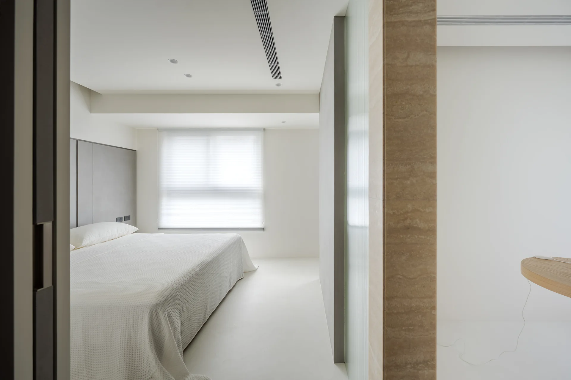 A minimalist white bedroom featuring a bed with white bedding, a gray paneled headboard, and a textured tan stone partition wall.