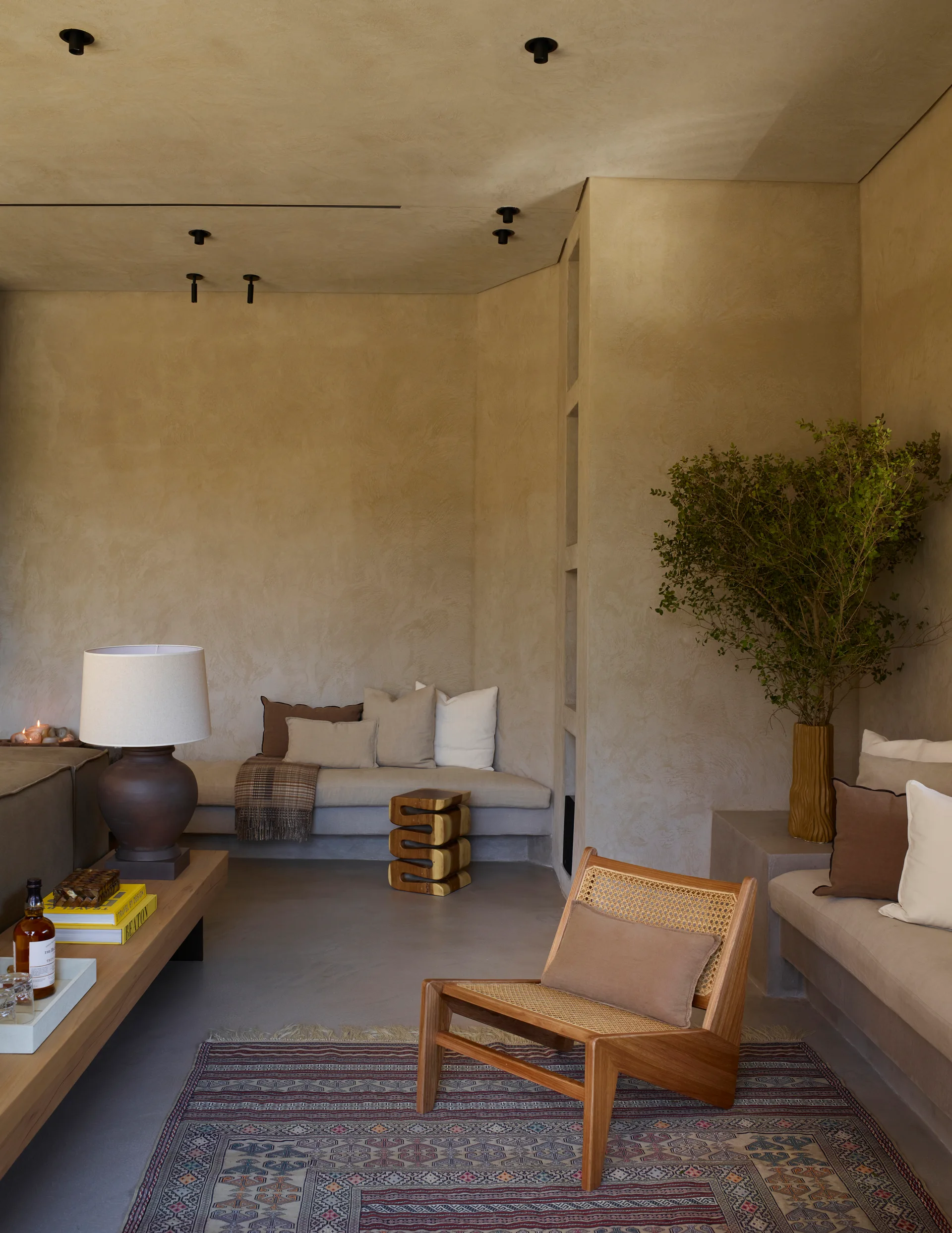 A plaster-walled lounge features built-in bench seating with linen cushions, a wooden cane armchair, and a patterned geometric area rug.