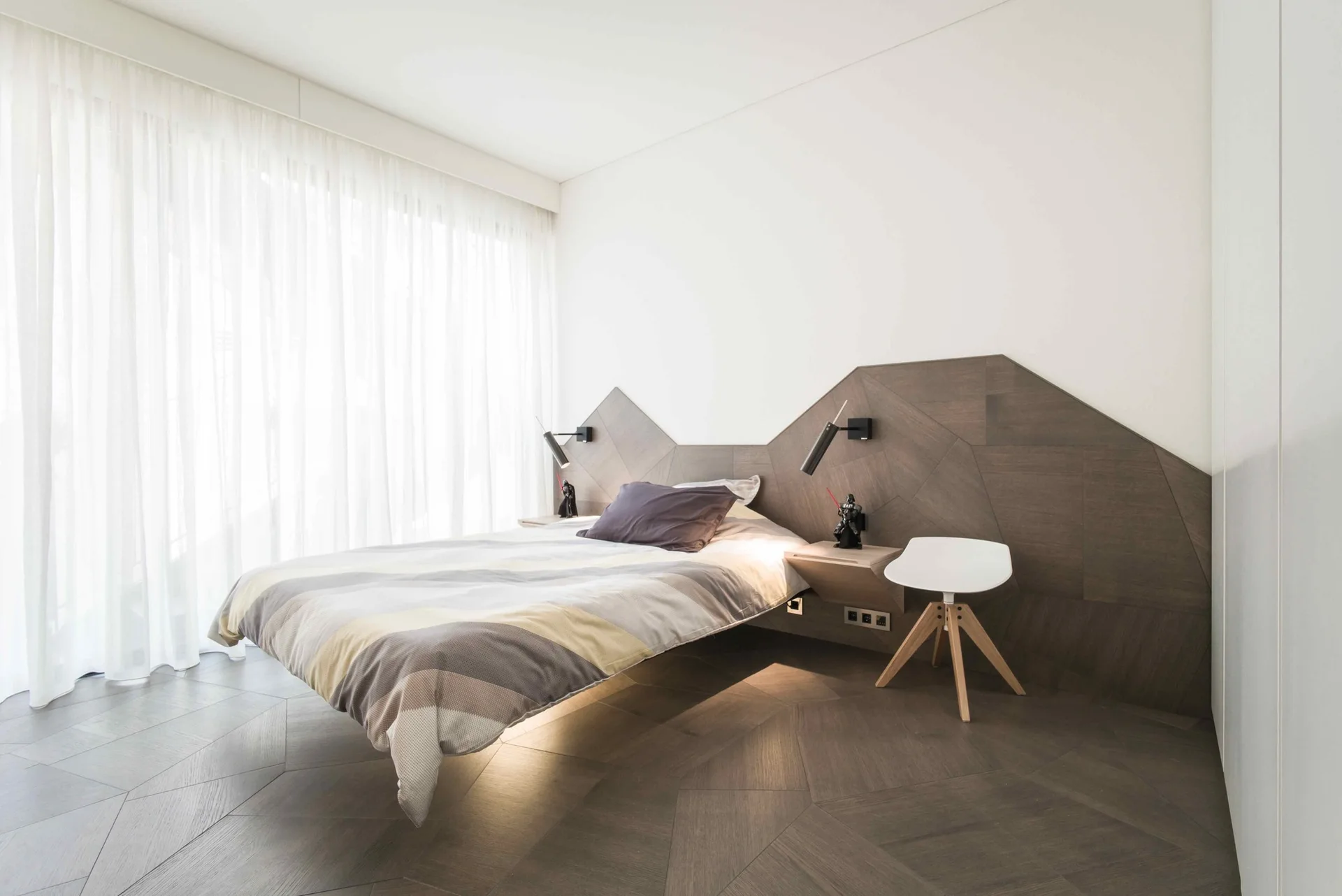 A modern bedroom features a low-profile bed against a custom geometric wood-paneled feature wall with floating nightstands and sheer white curtains.
