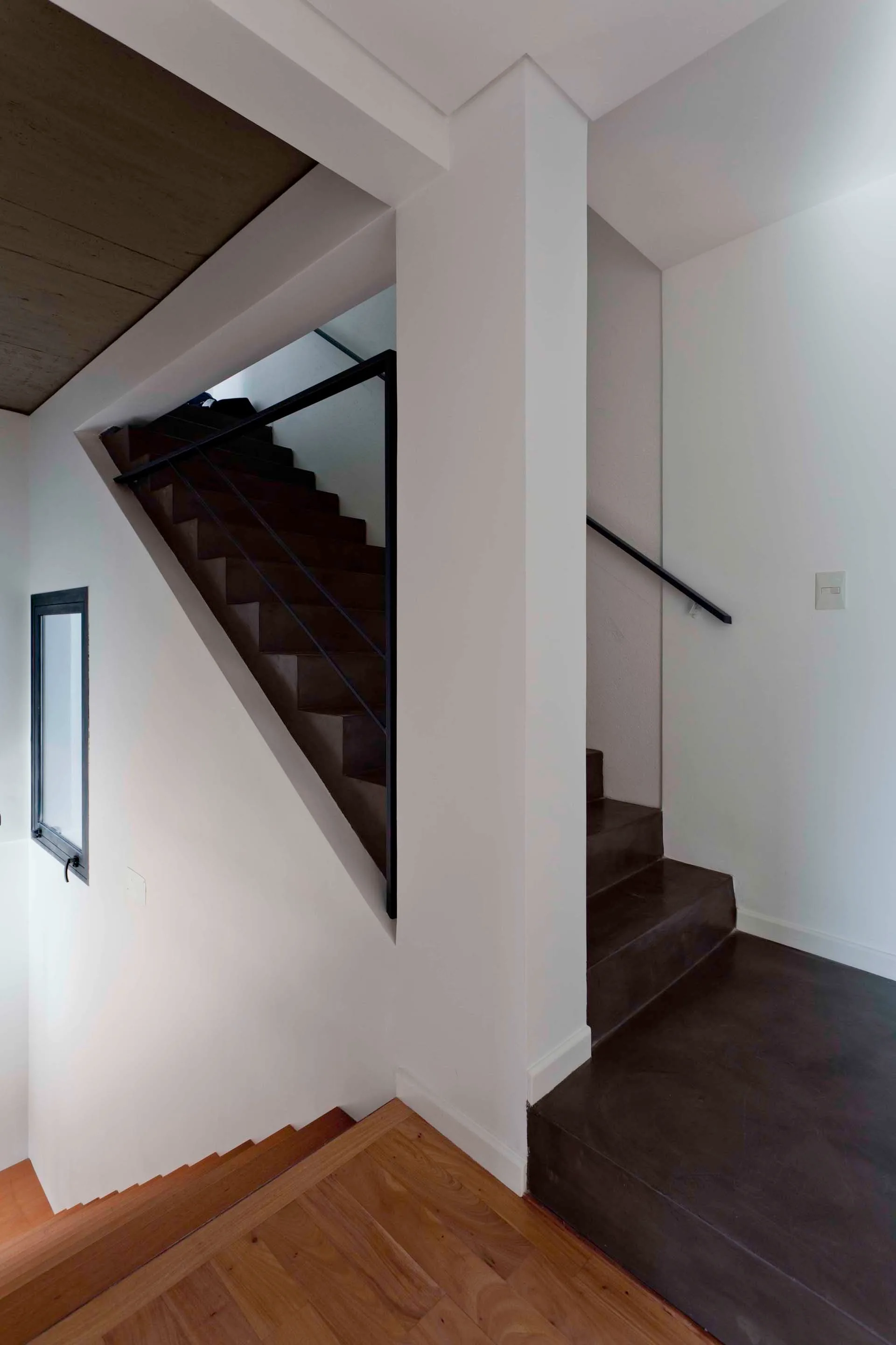 A minimalist multi-level staircase features sharp white walls, dark concrete treads, thin black metal railings, and natural wood steps.