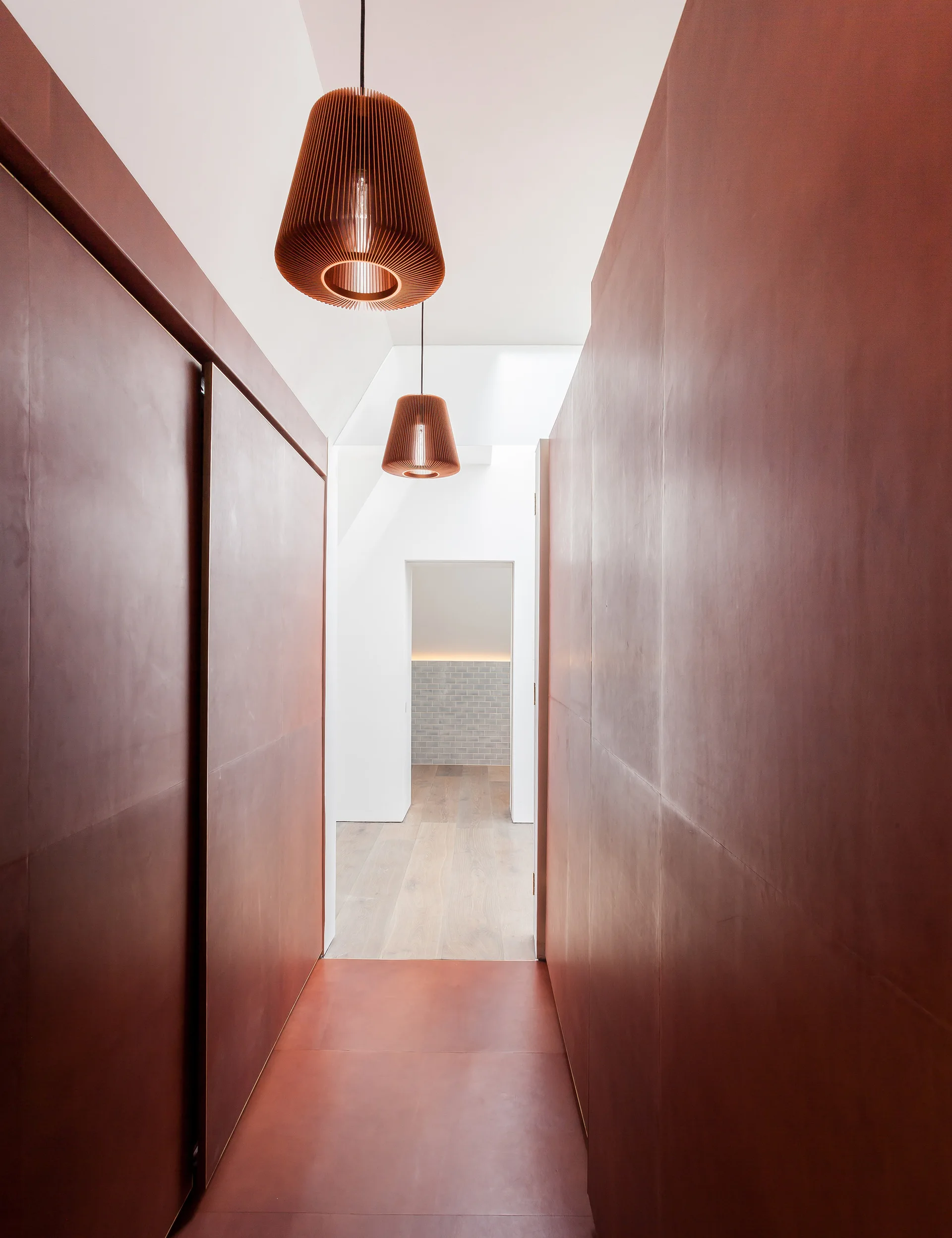 A modern hallway with floor-to-ceiling terracotta red wall panels, matching flooring, and two ribbed wooden pendant lights hanging from a white ceiling.