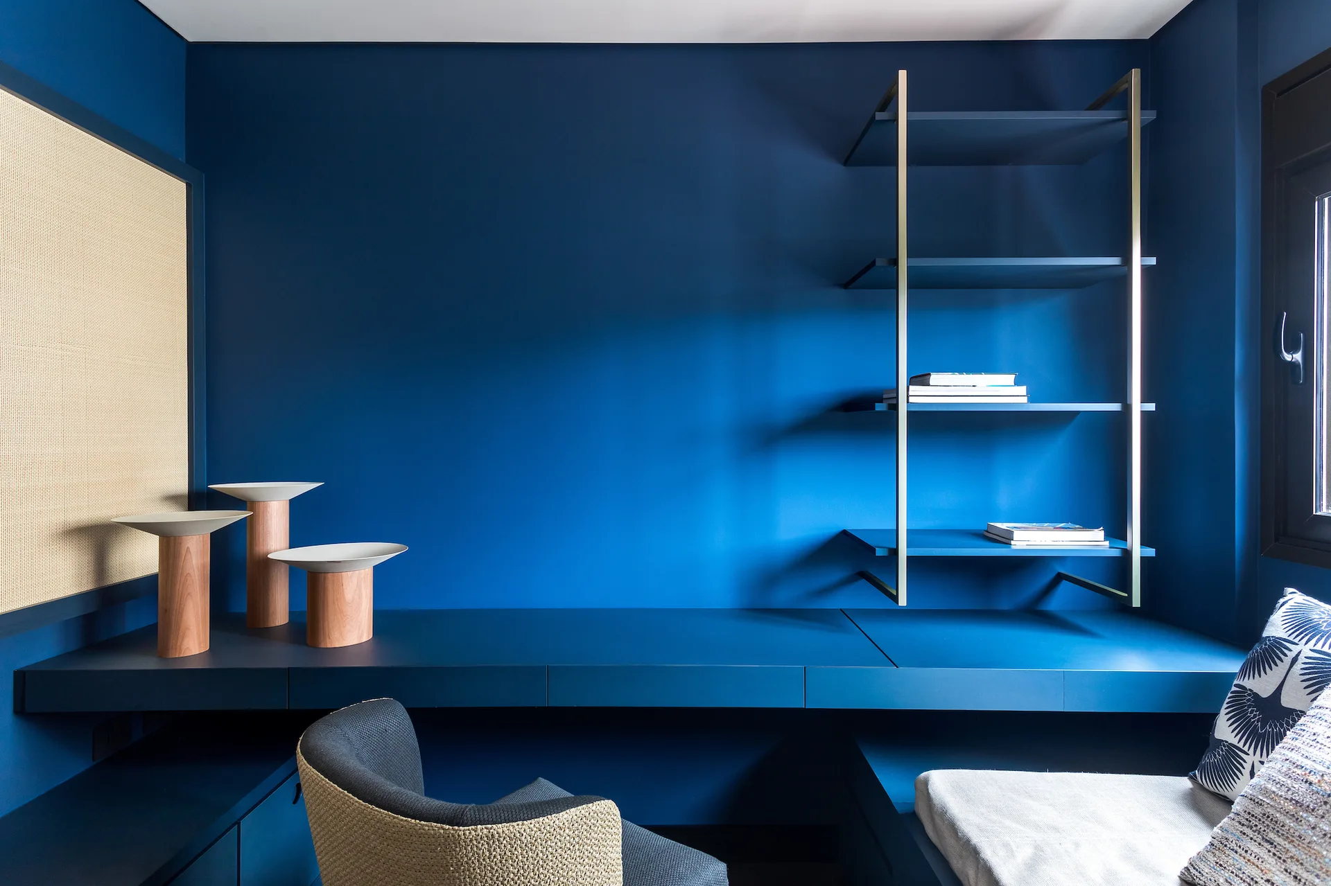 A minimalist home office features a monochromatic deep blue built-in desk, floating shelves, a woven cane wall panel, and a modern desk chair.