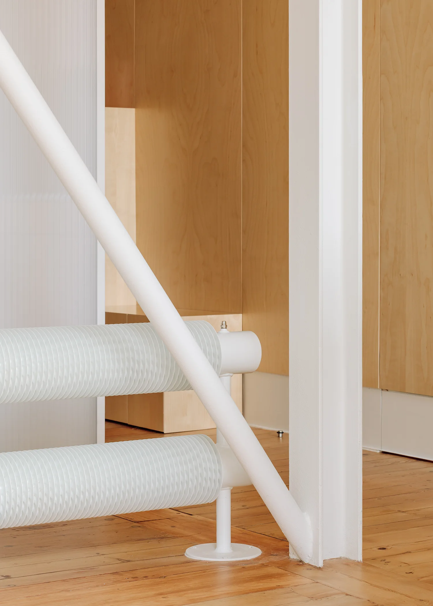 A diagonal white metal stair railing and cylindrical radiator set against warm plywood wall panels and light wood floors.