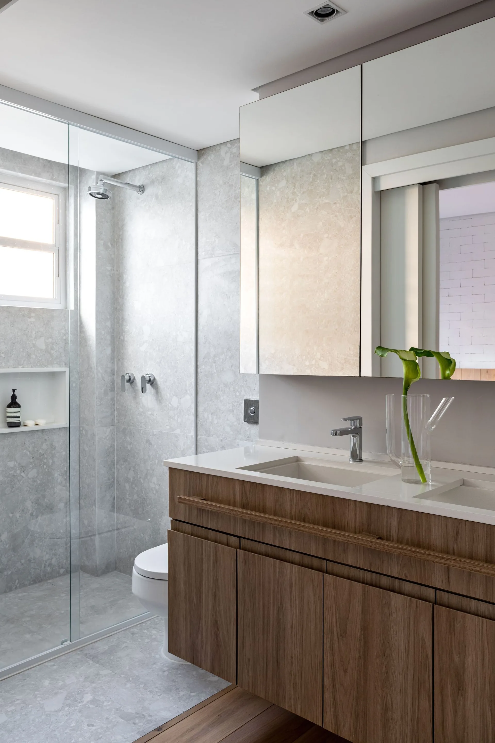 A modern bathroom showing a floating wood vanity with white sinks adjacent to a glass-enclosed shower tiled in grey speckled stone.